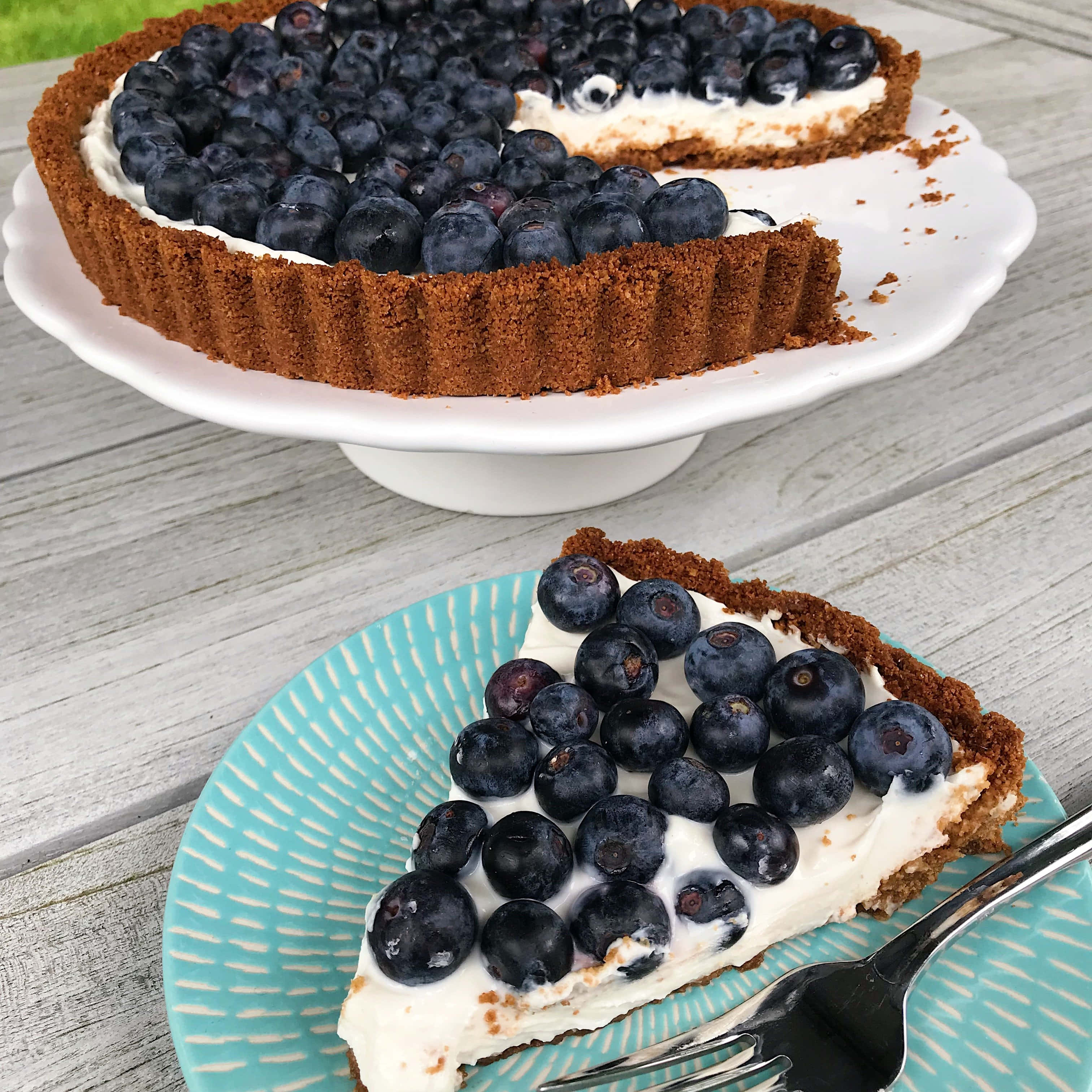 Blueberries Tart 桌布 壁紙 — 一塊藍莓塔放在青綠色盤子上，旁邊有一把叉子，其餘的塔則放在白色蛋糕架上，置於一張風化的木桌上。