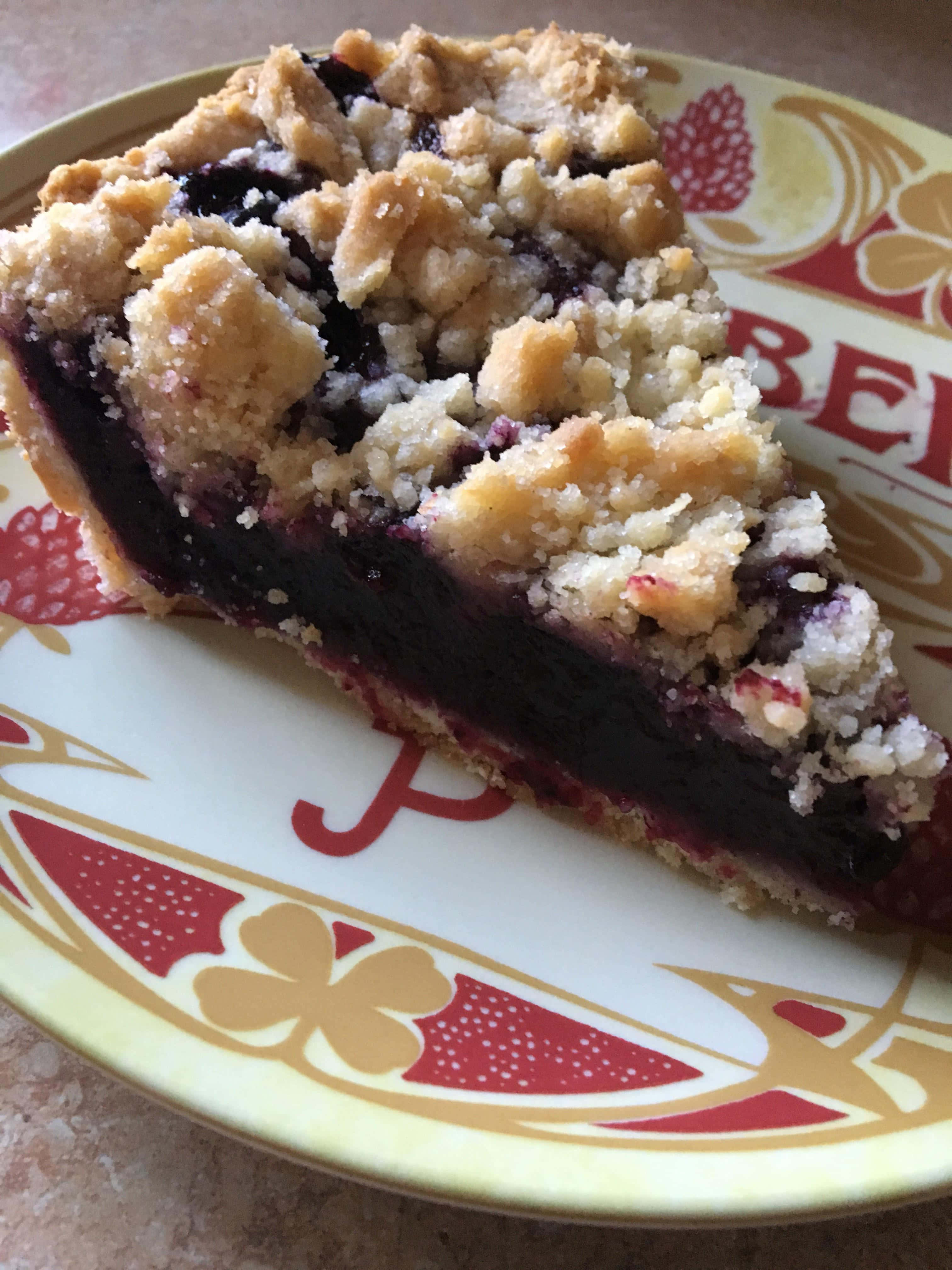 Blueberry Pie 桌布 壁紙 — 一塊藍莓金寶派的微距特寫，金黃酥皮放在有圖案的盤子上。