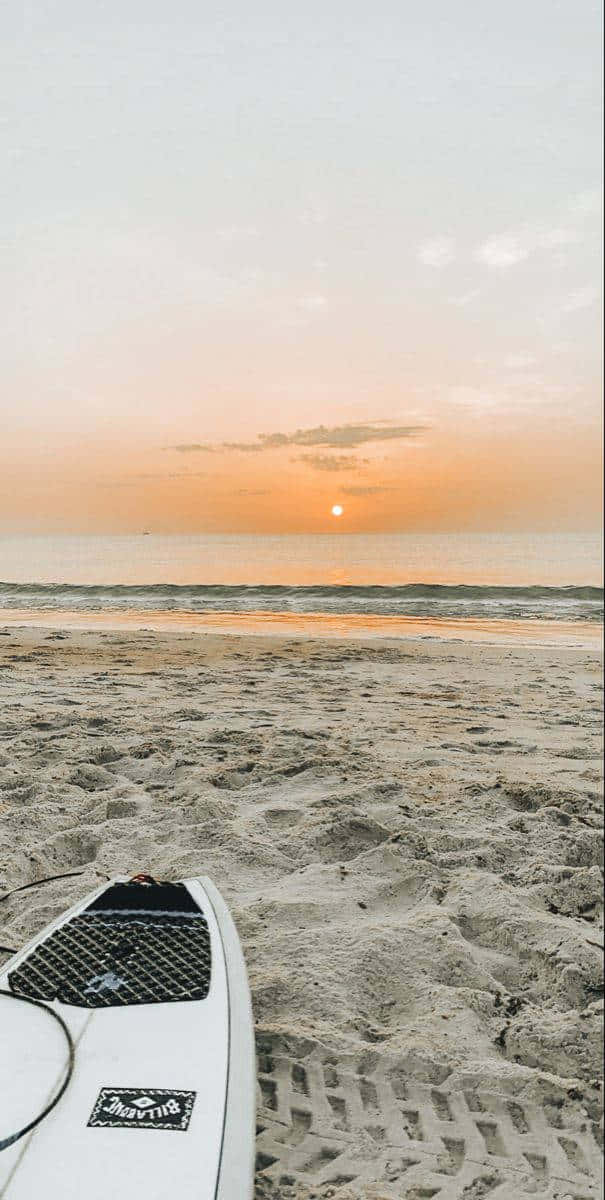 Sunset Beach 桌布 壁紙 — 一塊衝浪板擱在沙灘上，夕陽在平靜的海平線落下，散發出溫暖的橙色光芒。