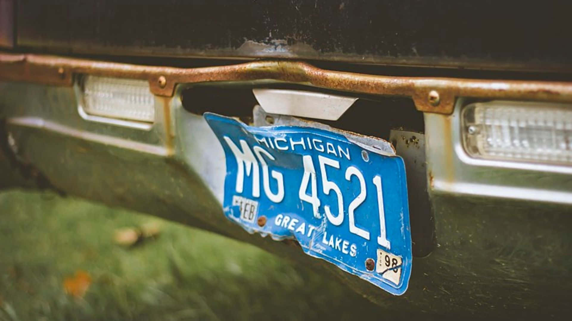 License Plate 桌布 壁紙 — 一塊褪色的藍色密西根州車牌，上面有「GREAT LAKES」字樣，歪斜地掛在生鏽的汽車保險桿上。