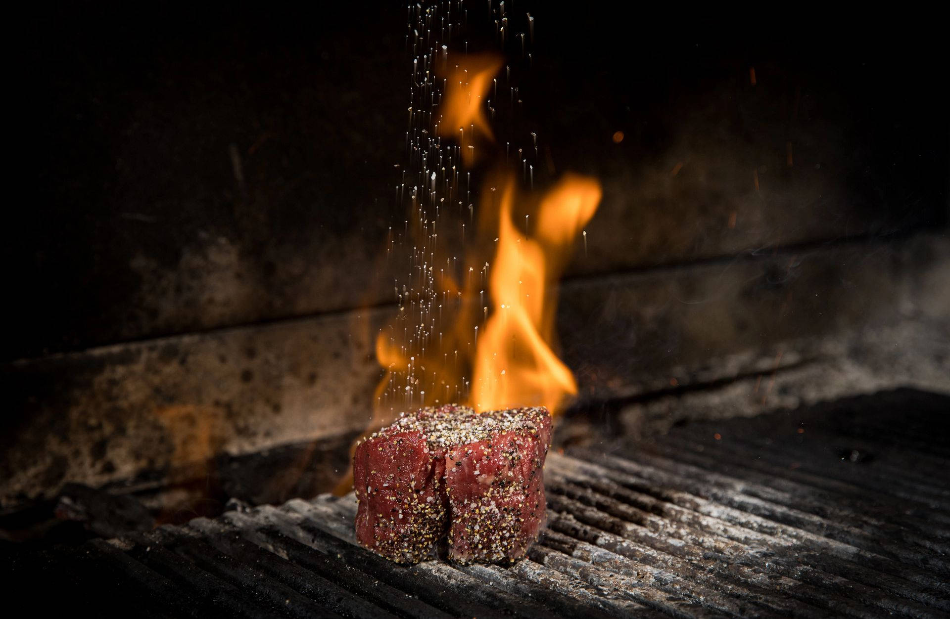 meat 桌布 壁紙 — 一塊調味牛排在烤架上滋滋作響，火焰躍起，鹽晶如瀑布般落下，形成戲劇性的特寫鏡頭。
