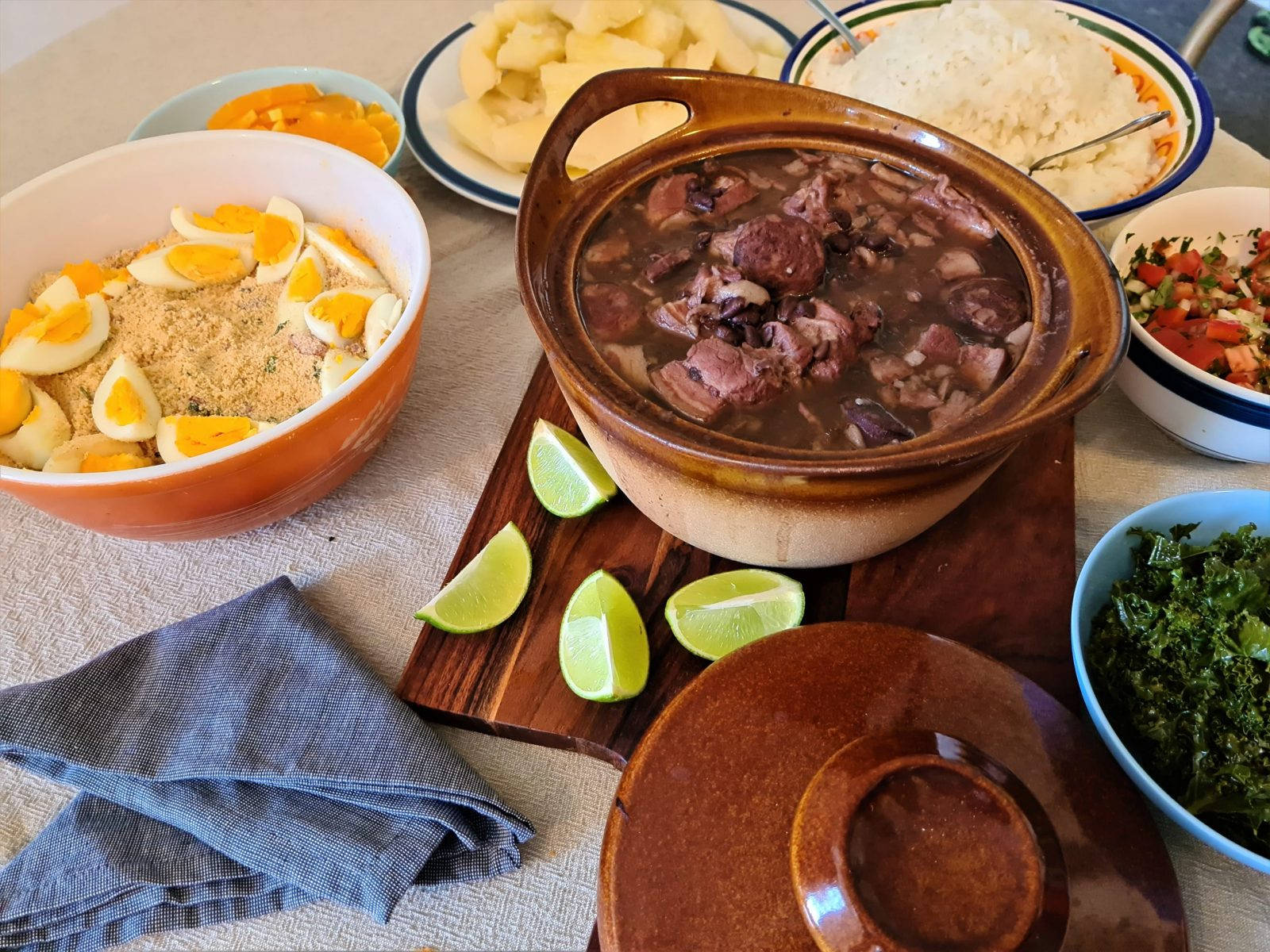 Delicious Food 桌布 壁紙 — 一塊質樸的木板上放著一個冒著熱氣的陶鍋，裡面是濃郁的深色燉菜，周圍環繞著色彩繽紛的配菜和萊姆角。