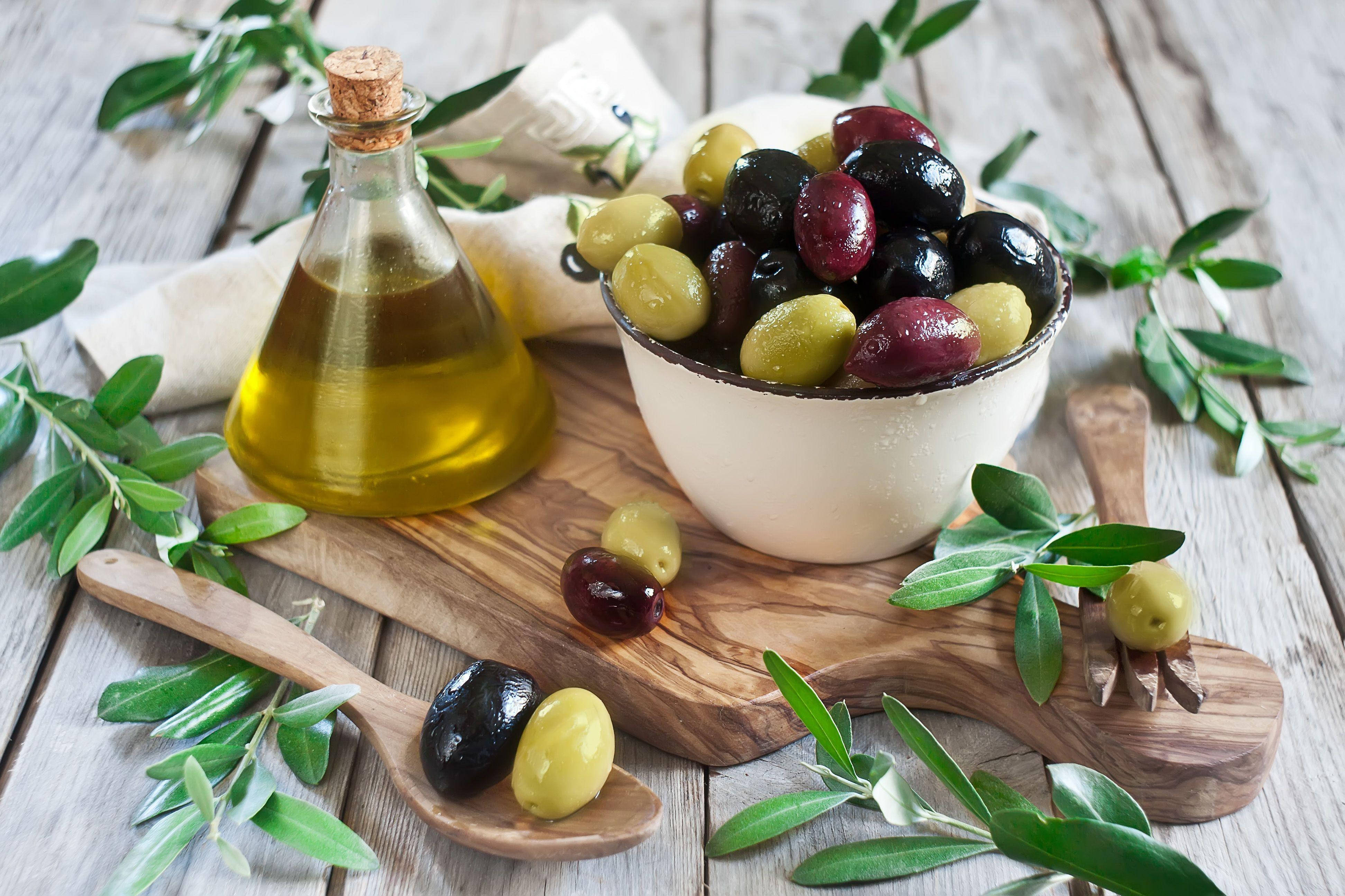 Olive 桌布 壁紙 — 一塊質樸的木板上放著一碗黑綠橄欖、一瓶金黃色的橄欖油，以及散落的橄欖葉，置於風化的木桌上。