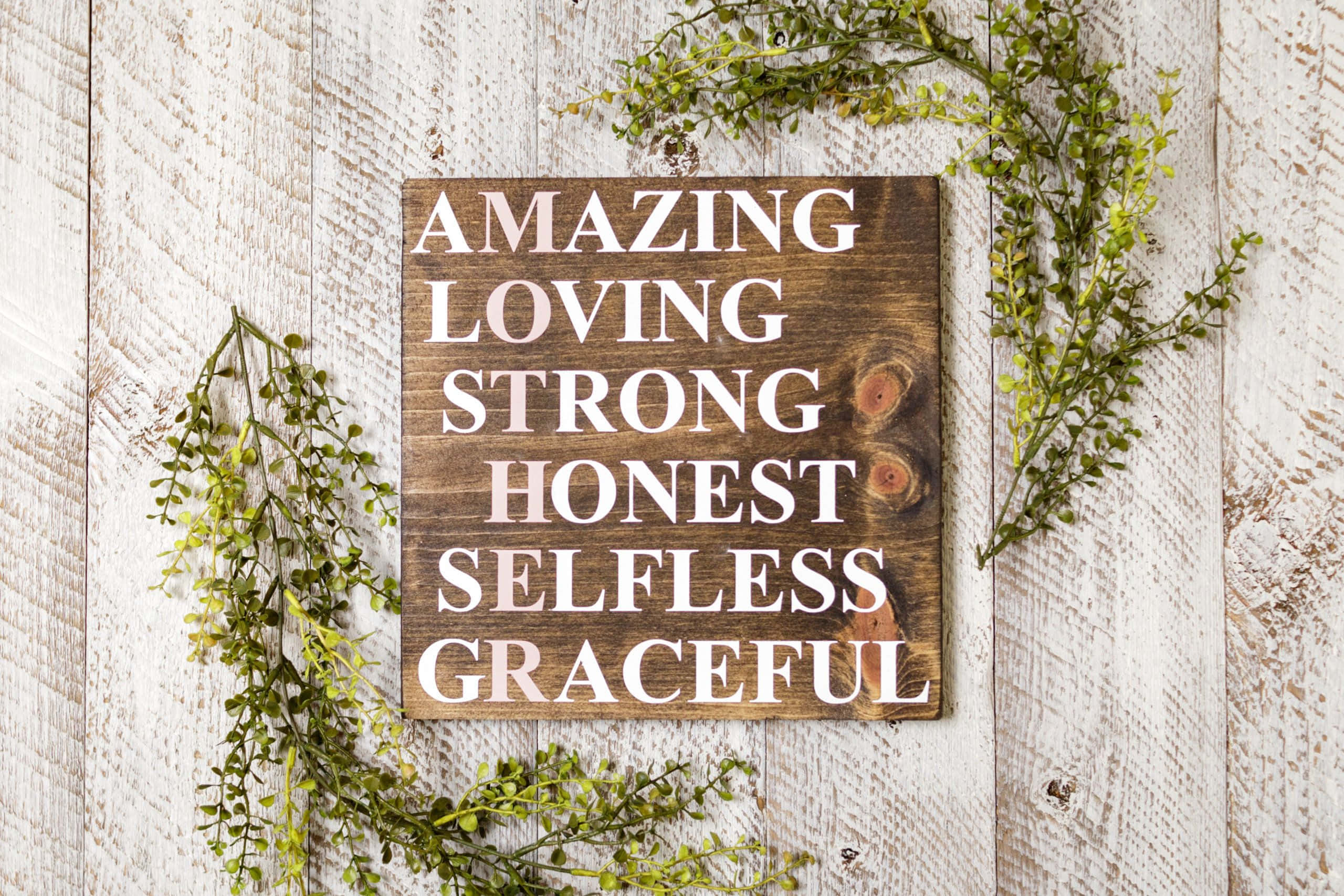 Woman 桌布 壁紙 — 一塊質樸的木牌，上面有白色文字寫著 amazing loving strong honest selfless graceful，周圍環繞著綠色植物，背景是風化的白色木材。