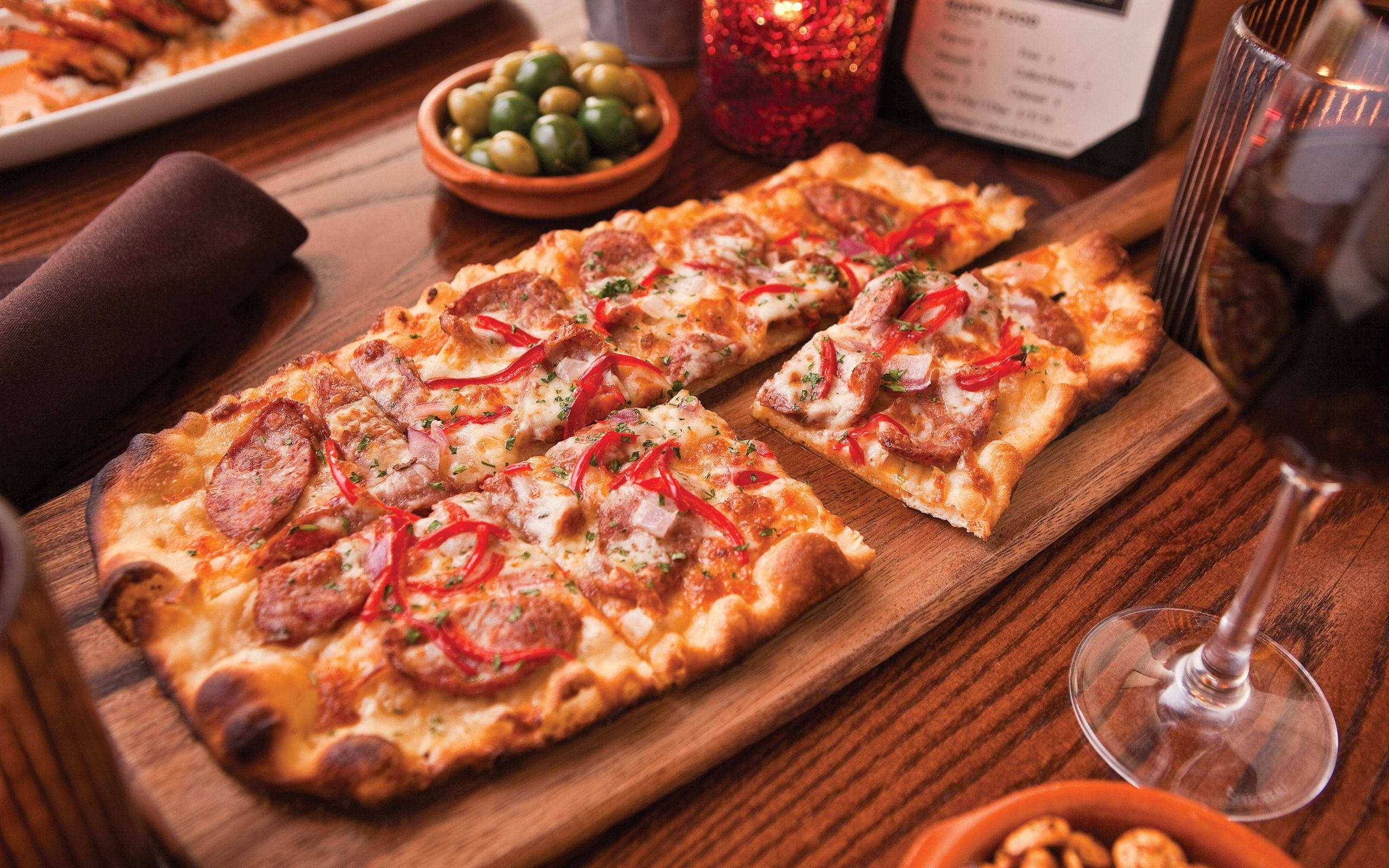 Pizza 桌布 壁紙 — 一塊鄉村風格的長方形披薩，上面鋪有義大利辣香腸、紅椒和起司，放在木板上，旁邊有一杯紅酒和橄欖。