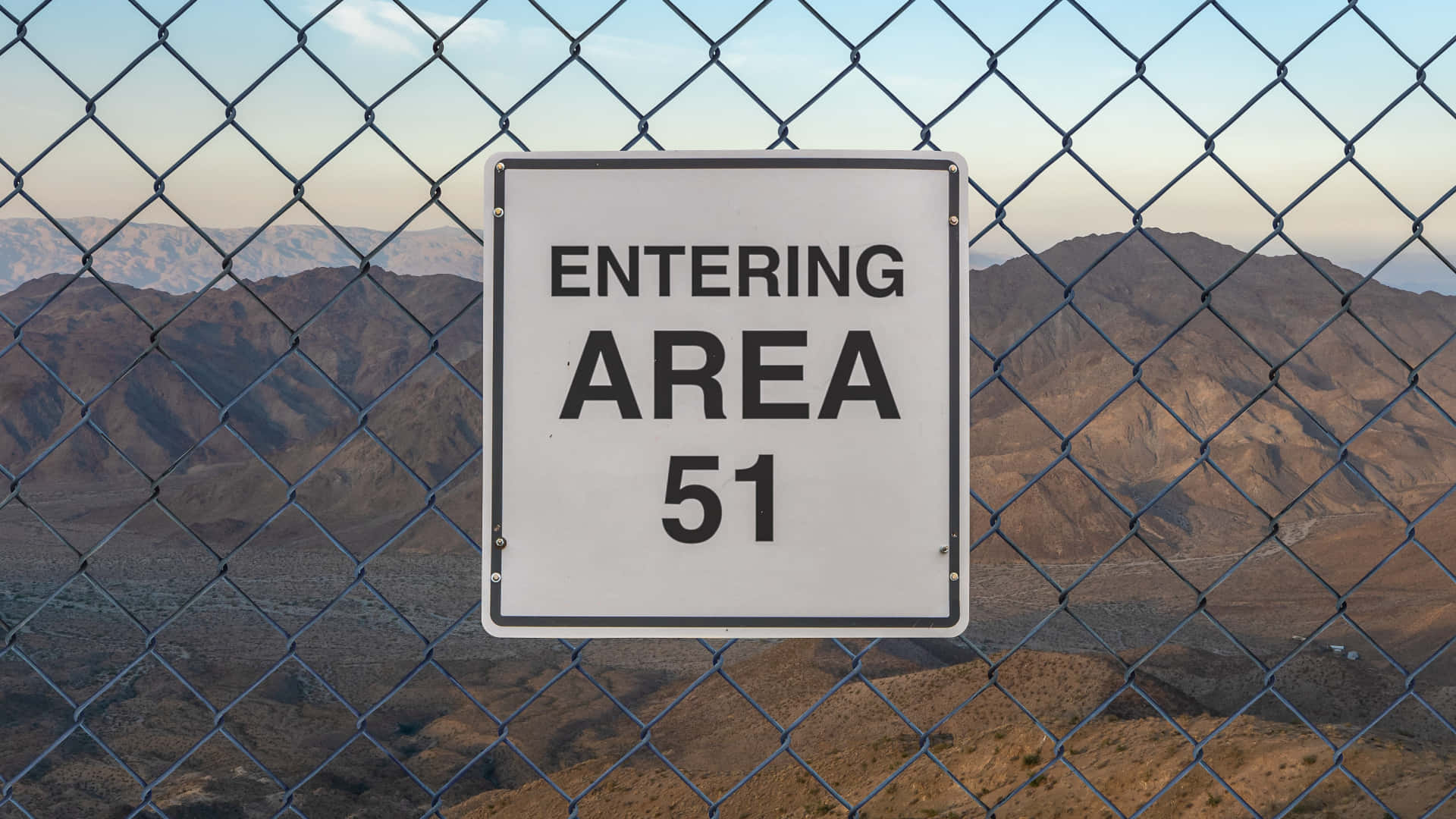 桌布 壁紙 — 一塊醒目的白色「ENTERING AREA 51」標誌掛在鐵絲網圍欄上，俯瞰著黃昏時分廣闊的沙漠景觀。