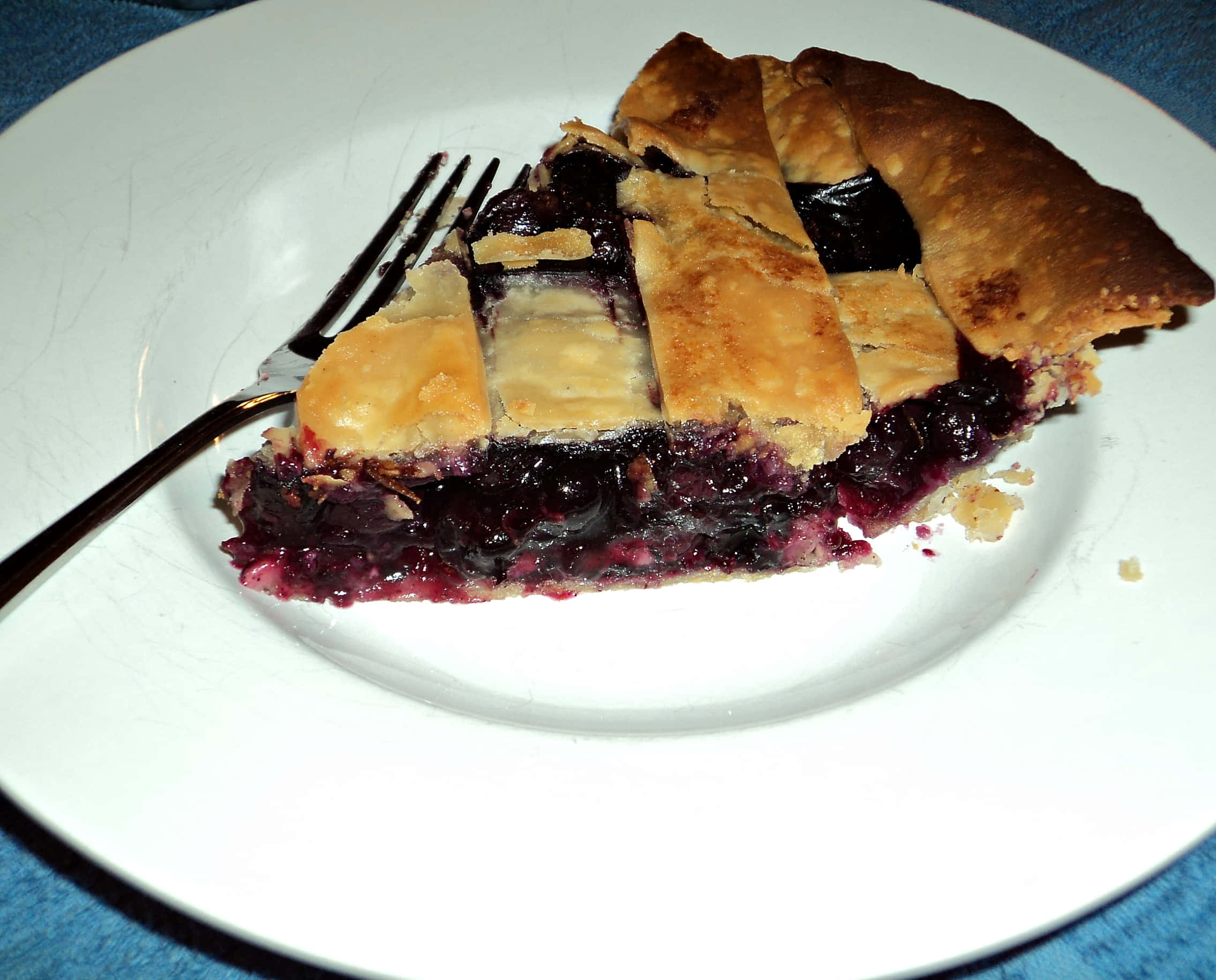 Blueberry Pie 桌布 壁紙 — 一塊金黃色格子頂藍莓派，內餡多汁，放在白色盤子上，附有叉子。