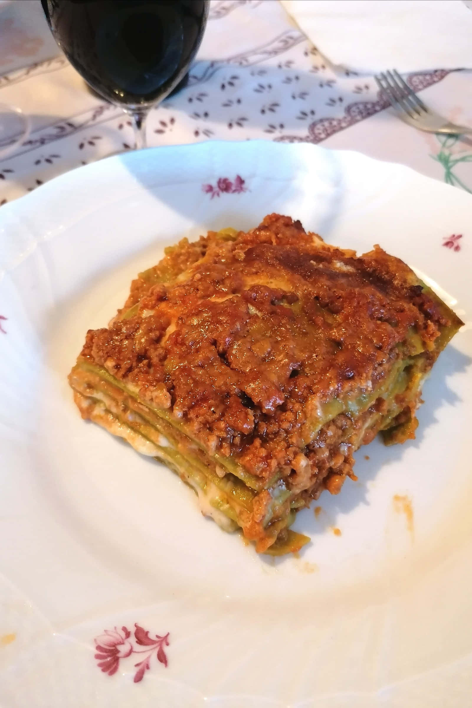meat 桌布 壁紙 — 一塊金黃色的千層麵，配上濃郁的番茄醬和融化的起司，放在有紅色花卉圖案的白色盤子上。