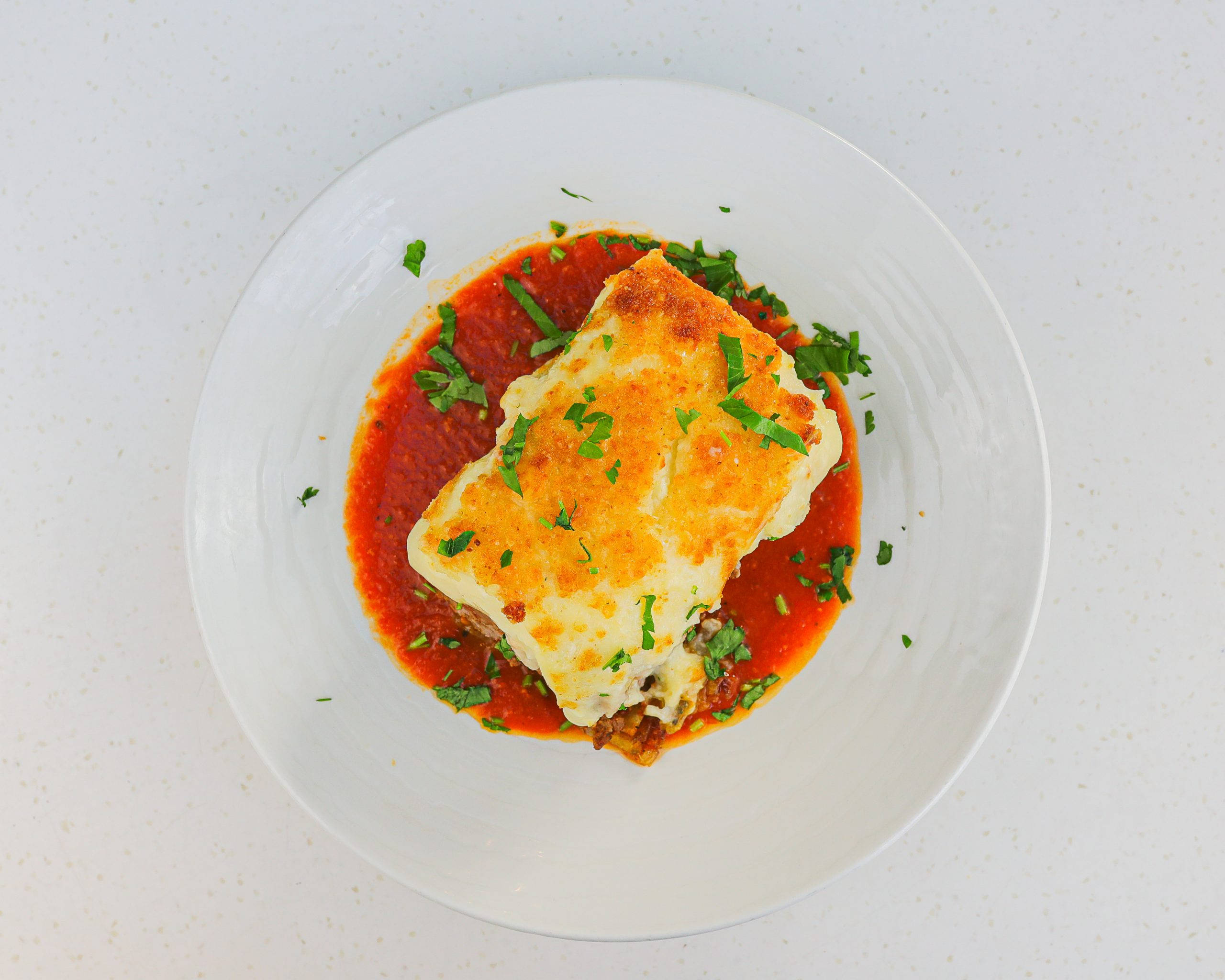 Food Photography 桌布 壁紙 — 一塊金黃色的千層麵放在濃郁的番茄醬中，置於白色盤子上，並以新鮮的綠色香草裝飾。