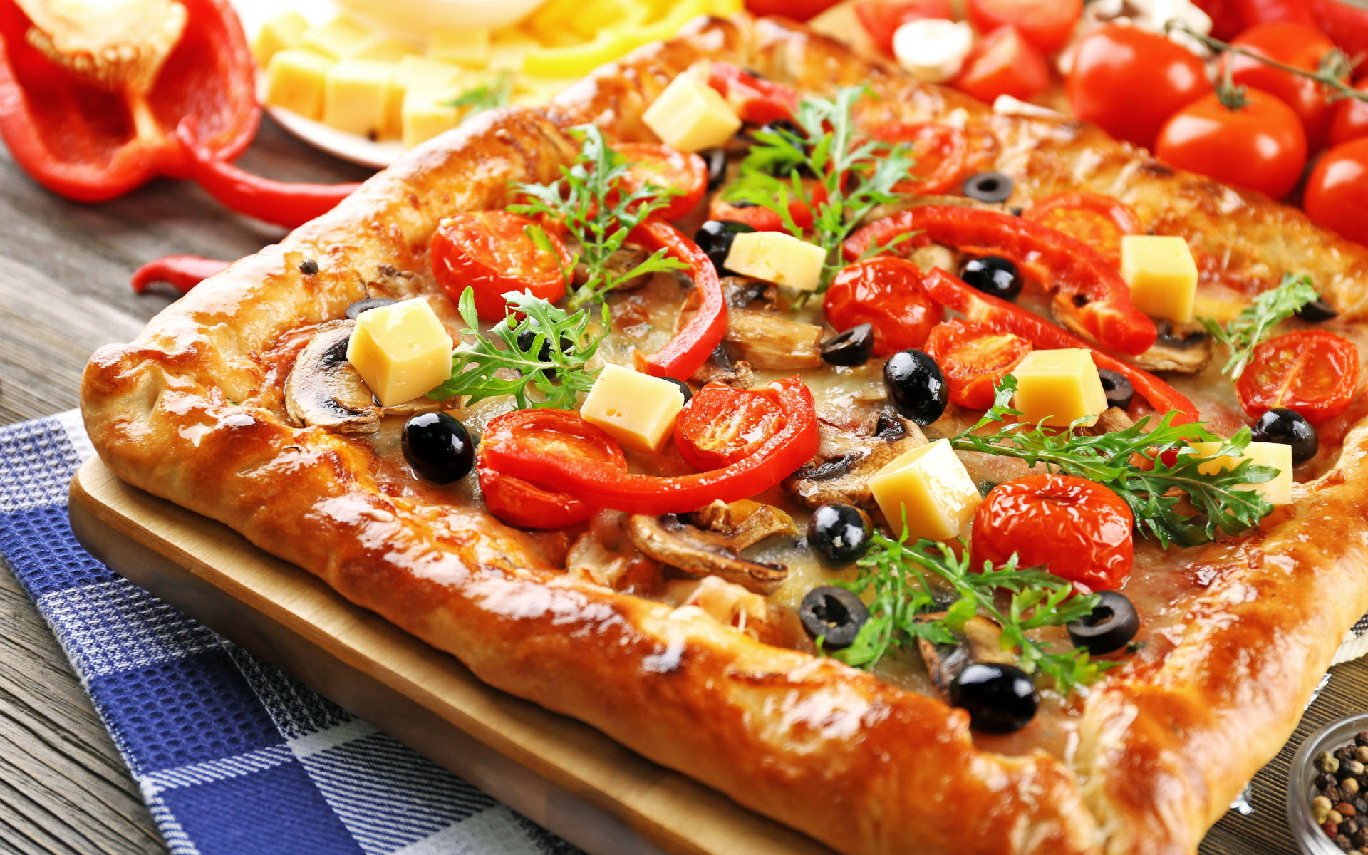 Pizza 桌布 壁紙 — 一塊金黃色的長方形披薩，上面鋪滿番茄、青椒、橄欖和起司，放在藍色格子布上的木板上。