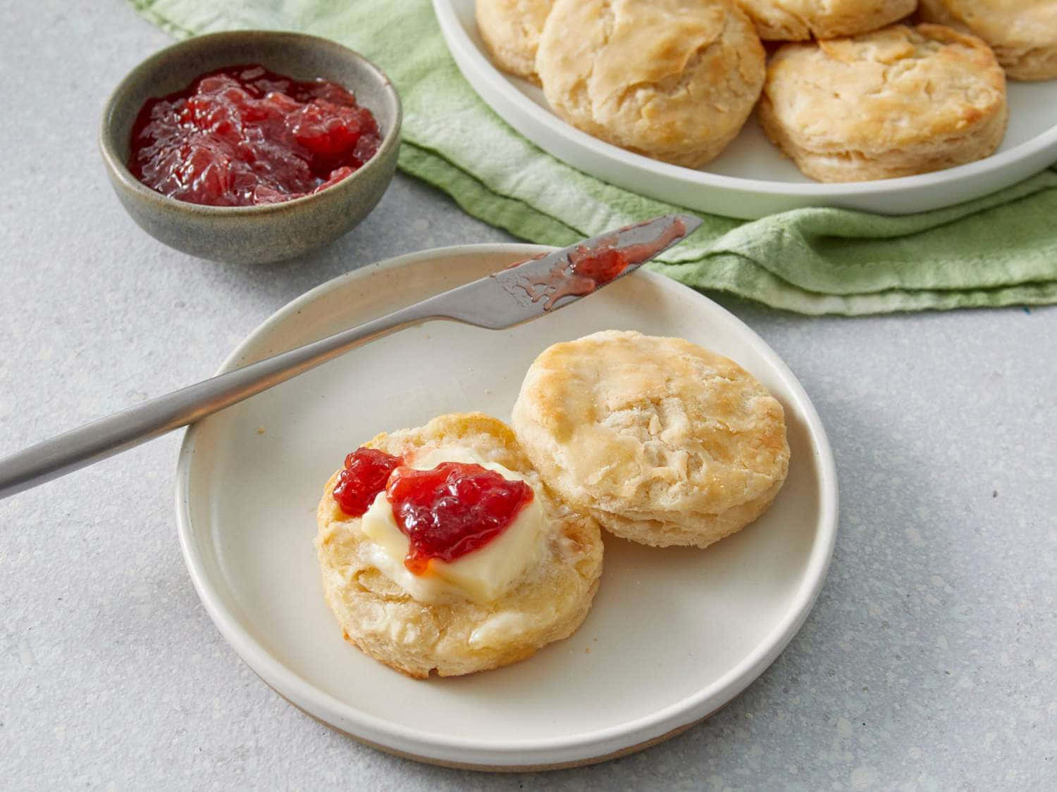 Biscuit 桌布 壁紙 — 一塊金黃色的餅乾被掰開，上面有奶油般的奶油和鮮紅色的果醬，放在白色盤子上，旁邊有一把刀和更多餅乾。