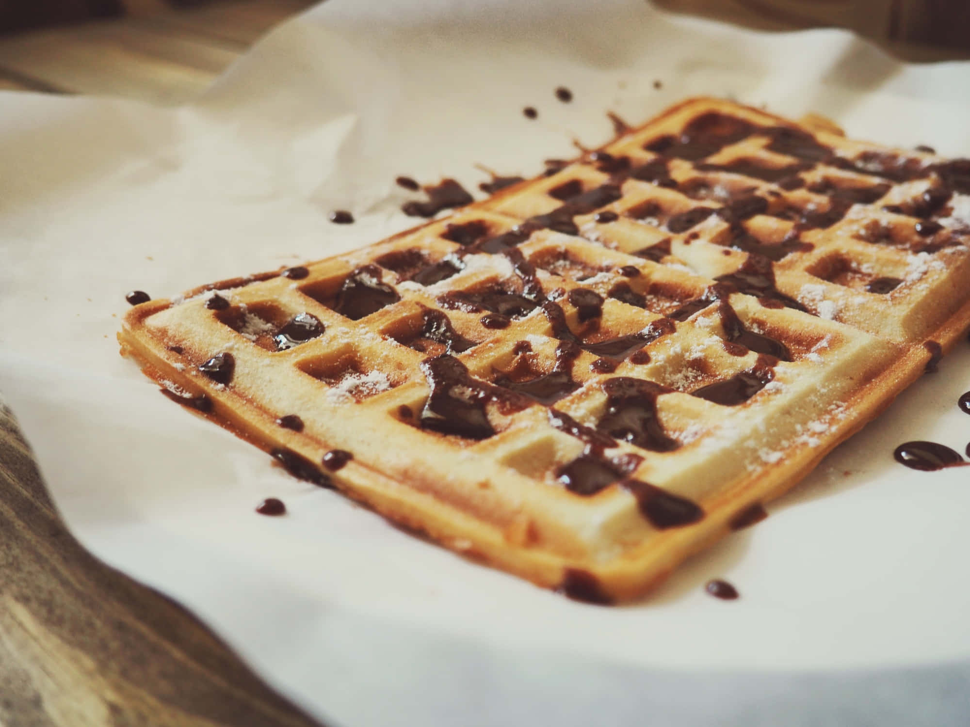 Waffle 桌布 壁紙 — 一塊金黃色的鬆餅淋上濃郁的巧克力醬並撒上糖粉，放在烘焙紙上。