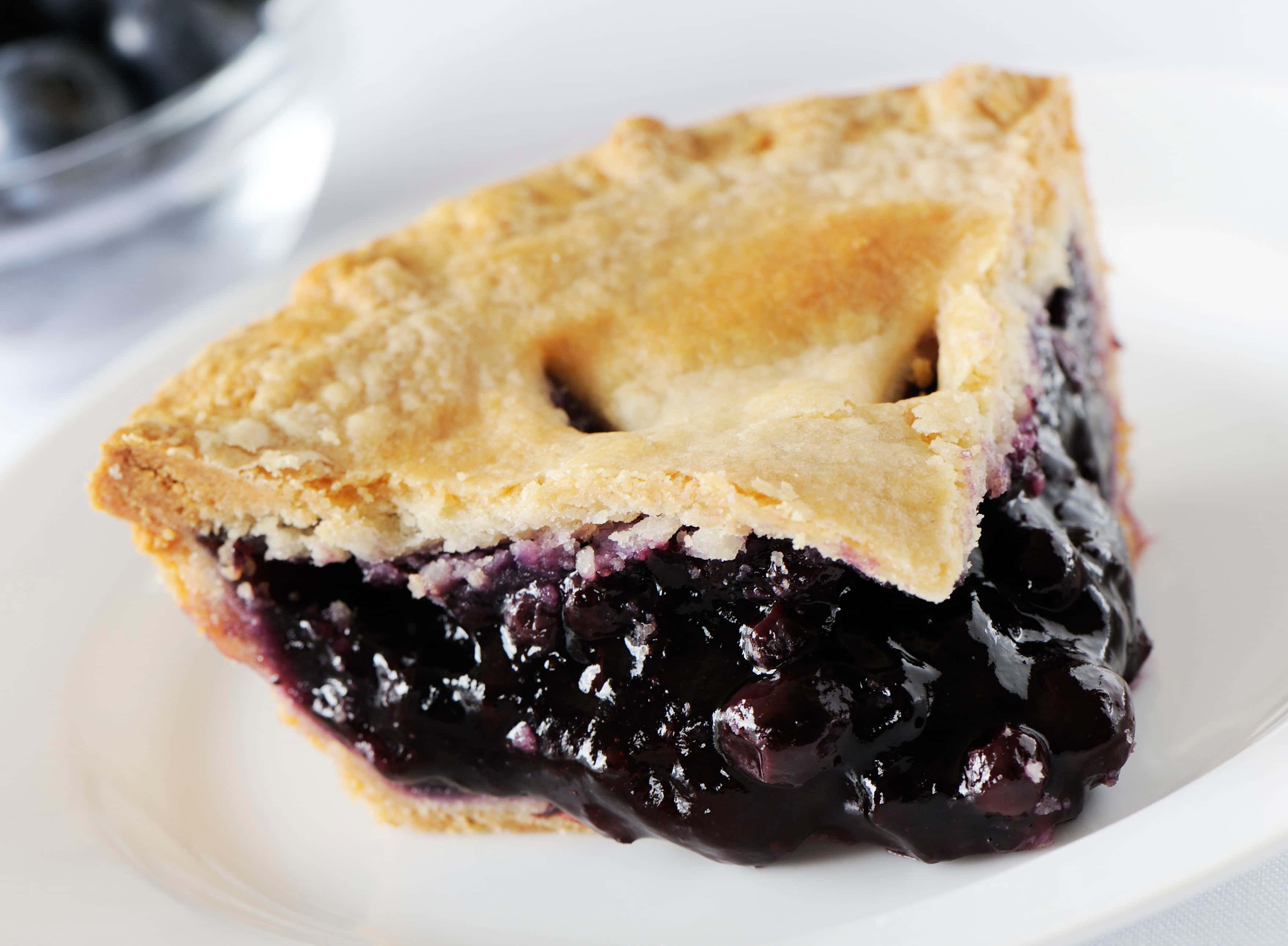Blueberry Pie 桌布 壁紙 — 一塊金黃酥脆的藍莓派，帶有 juicy 深色餡料，放在白色盤子上，背景有一碗新鮮莓果。