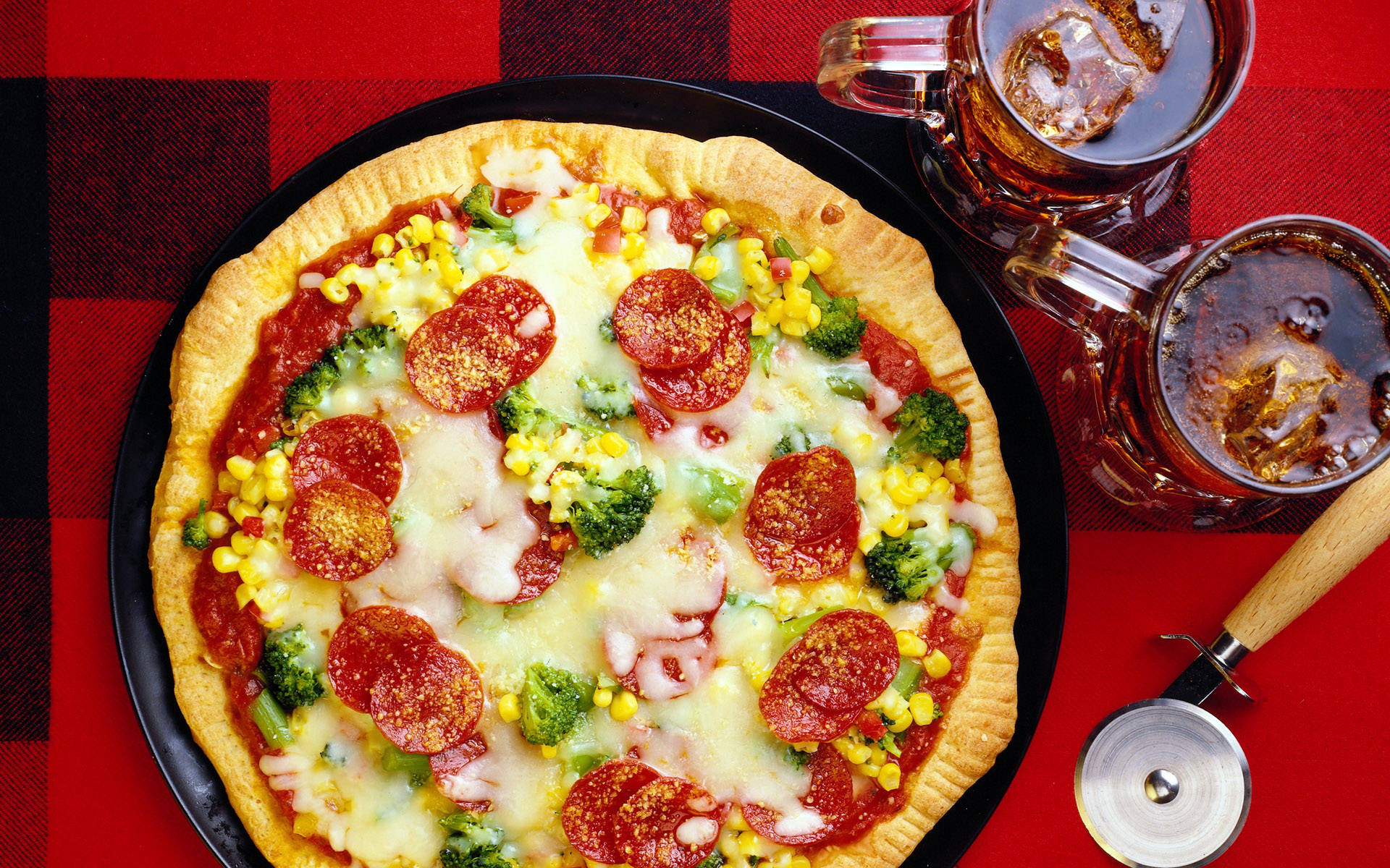 Pizza 桌布 壁紙 — 一塊鋪滿起司、辣肉腸、花椰菜和玉米的披薩放在黑色盤子上，旁邊有兩杯冰茶，置於紅色格紋桌布上。