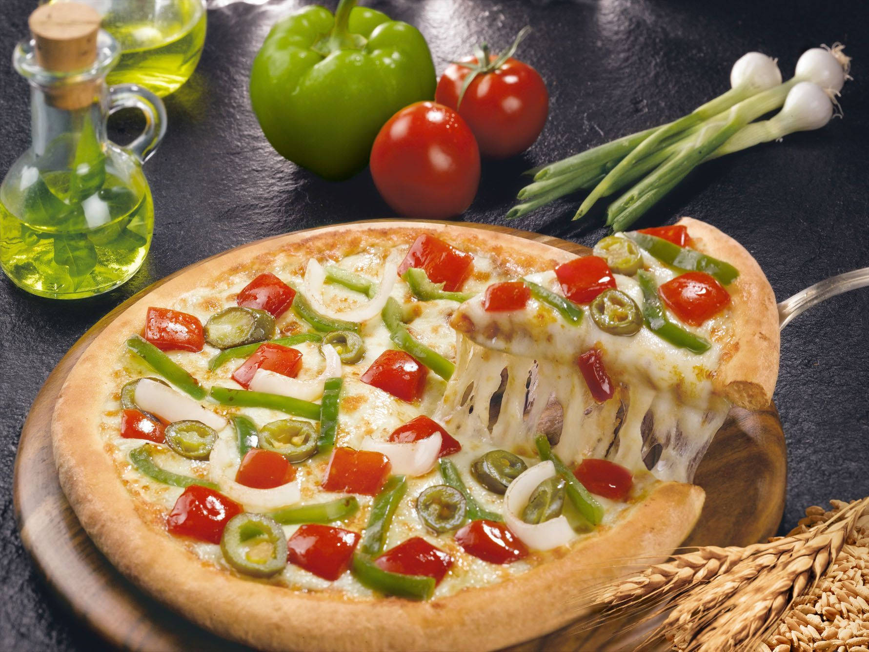 Dominos Pizza 桌布 壁紙 — 一塊鋪滿起司、青椒、洋蔥和番茄的披薩，正用鍋鏟在深色石板表面上鏟起，周圍放滿新鮮食材。