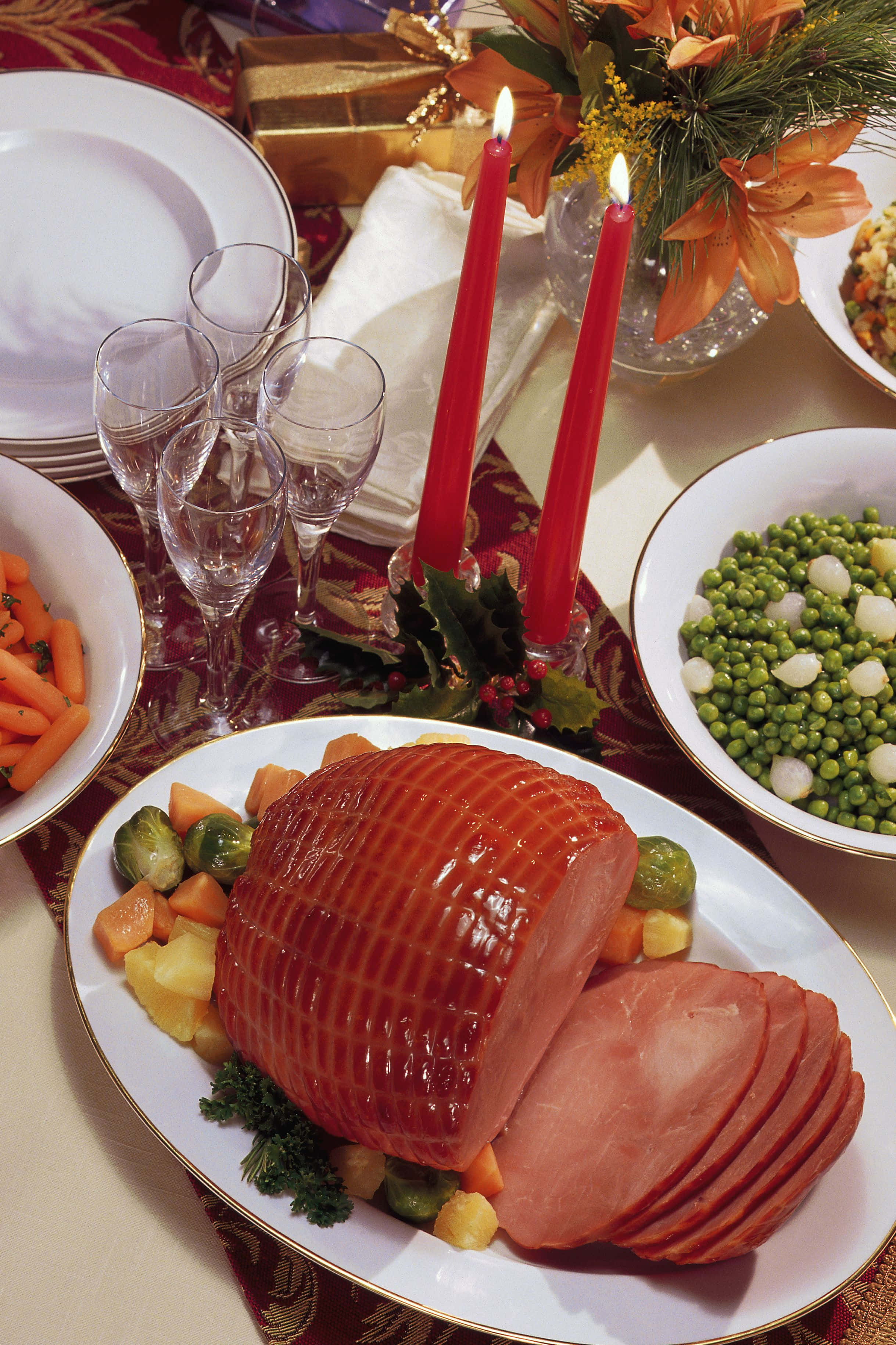Black Forest Ham 桌布 壁紙 — 一塊閃亮的烤火腿切片放在大盤上，周圍環繞著蔬菜和點燃的紅色蠟燭，擺在節日餐桌。