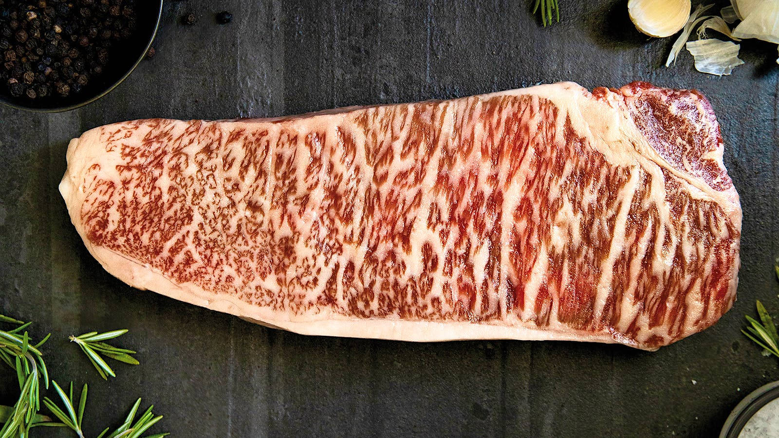 meat 桌布 壁紙 — 一塊頂級大理石紋牛排放在深色石板表面上，周圍環繞著新鮮迷迭香、胡椒粒和大蒜。