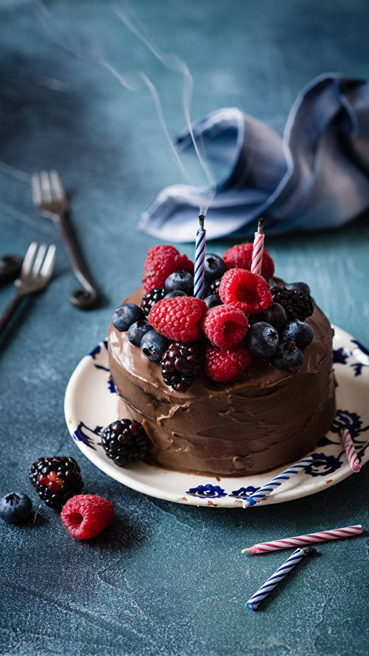 Birthday Cake 桌布 壁紙 — 一塊頹廢的巧克力蛋糕，上面鋪滿新鮮莓果和冒煙的蠟燭，放在一個有圖案的盤子上，背景是深沉的青綠色。