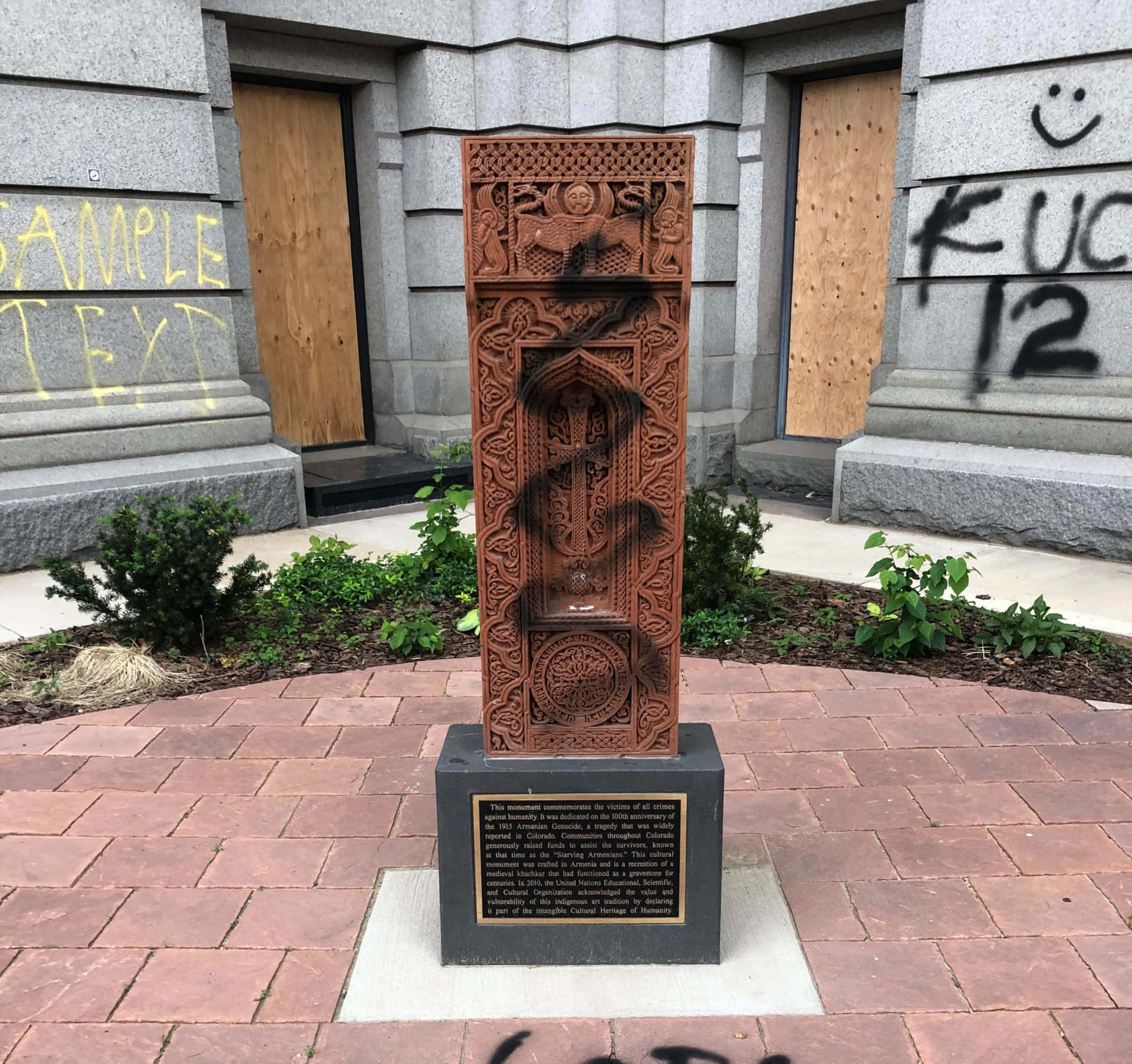 Memorial 桌布 壁紙 — 一塊風化、華麗的紅色紀念牌豎立在石基座上，前方是一座灰色石建築，門被封板封住，並有塗鴉。