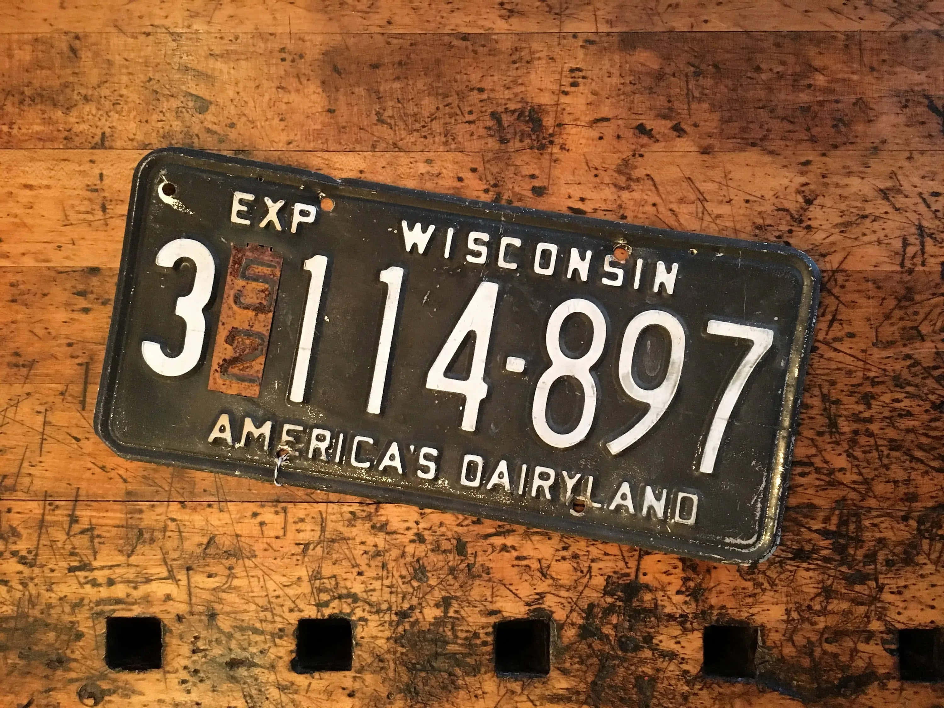 License Plate 桌布 壁紙 — 一塊風化的威斯康辛州車牌，上面有'America's Dairyland'字樣，擱在刮傷的木質表面上。