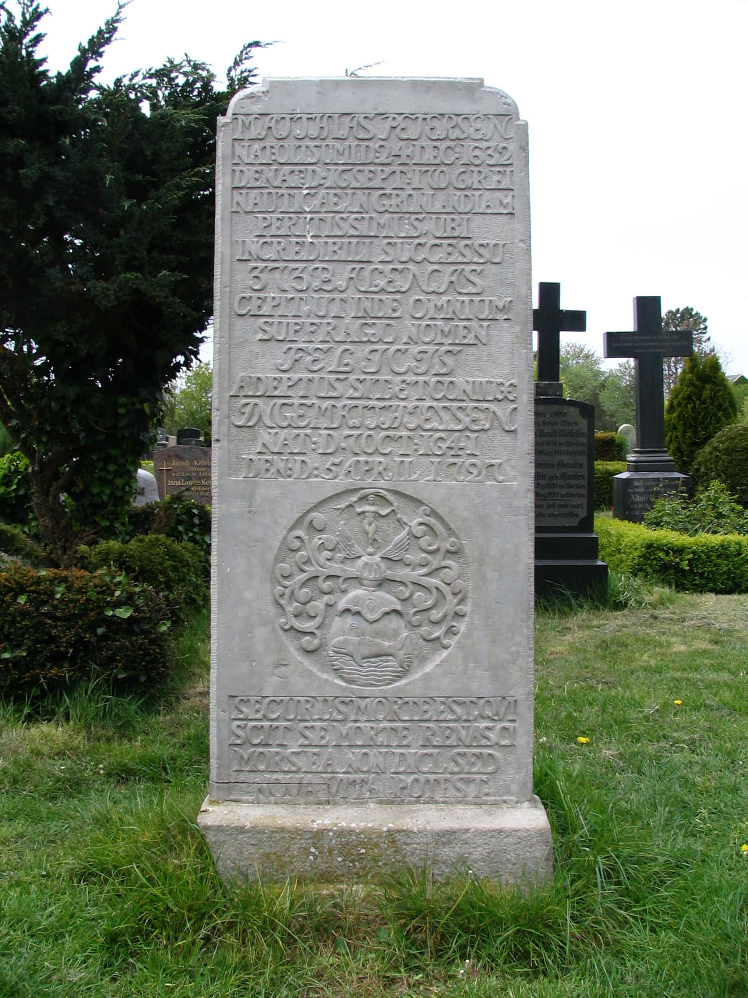 Gravestone 桌布 壁紙 — 一塊風化的灰色石墓碑，上面刻有文字和圓形徽章，矗立在一個被樹木和其他墓碑環繞的草地墓園中。