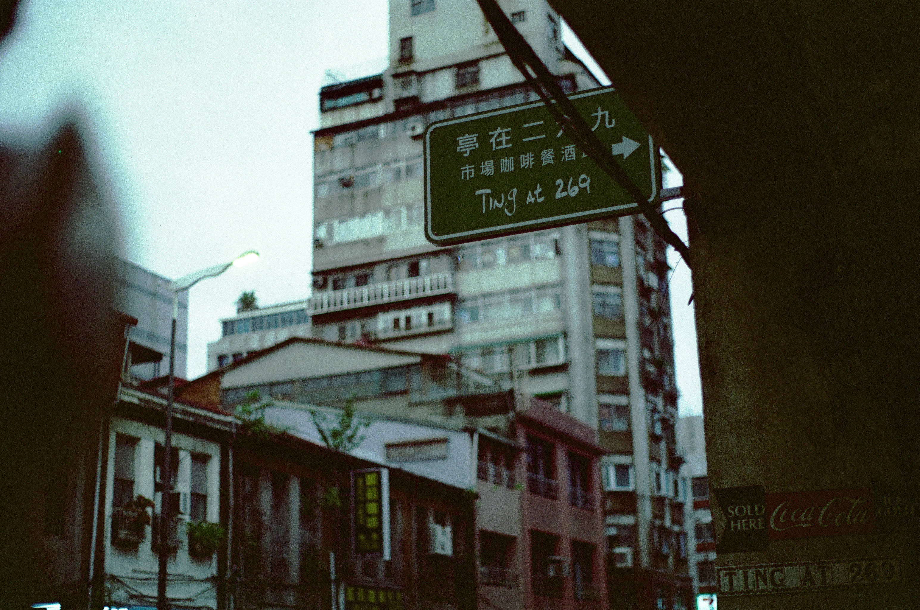 美學攝影 桌布 壁紙 — 一塊風化的綠色路牌，上面有手寫文字，掛在陰暗的城市小巷中，兩旁是高樓大廈，天空多雲。
