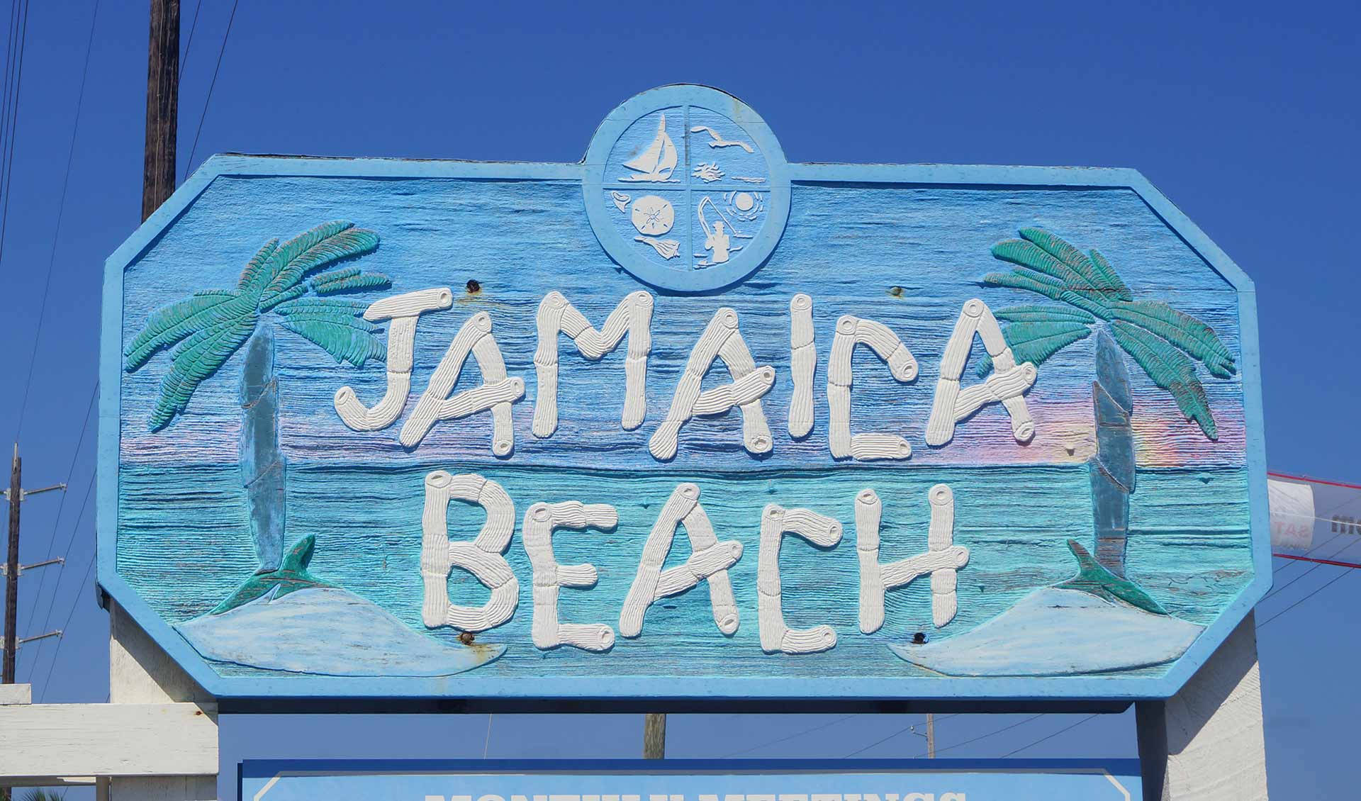 Jamaica Beach 桌布 壁紙 — 一塊風化的藍色木牌上寫著 Jamaica Beach，上面繪有棕櫚樹和海洋波浪，背景是晴朗的天空。