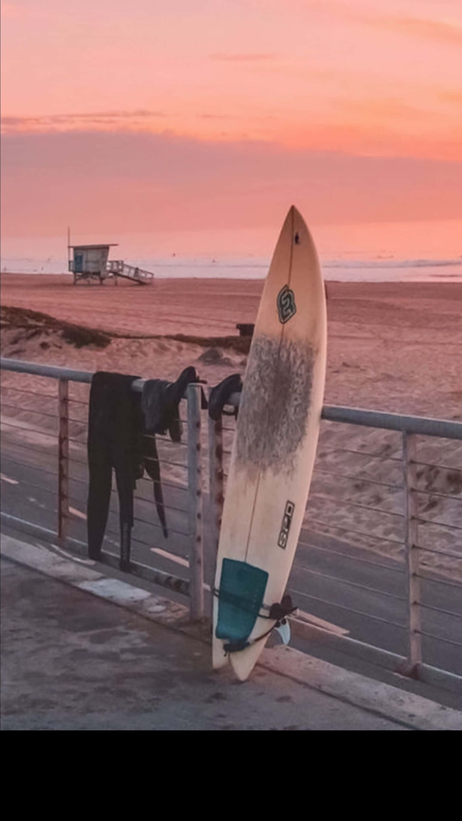 Beach Scene 桌布 壁紙 — 一塊風化的衝浪板在日落時靠在欄杆上，遠處有救生塔。