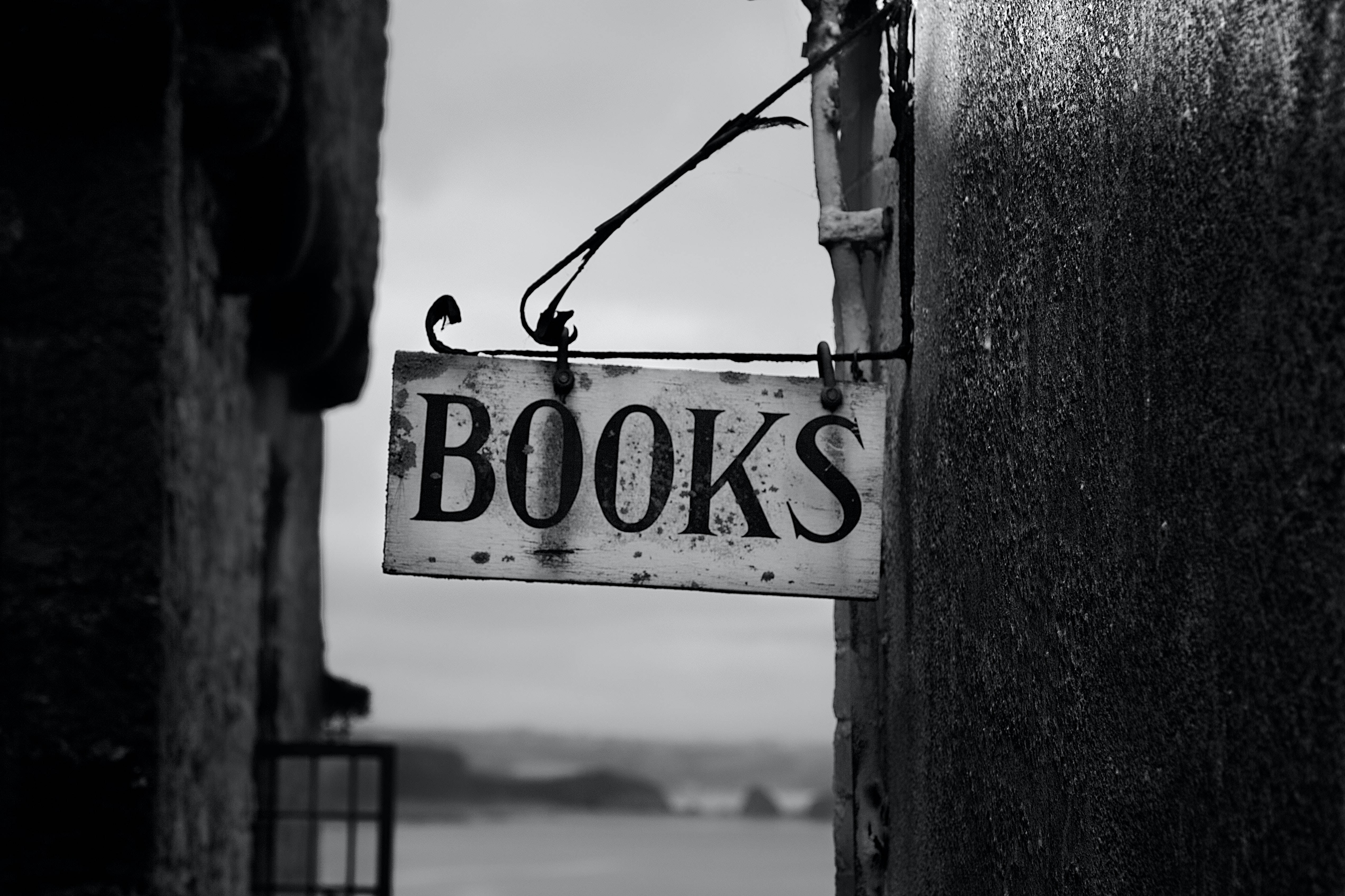 書本 桌布 壁紙 — 一塊風化的黑白「BOOKS」標誌掛在石牆之間，俯瞰霧氣瀰漫的海岸景色。