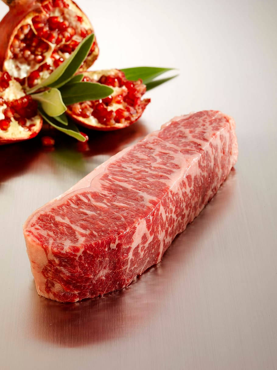 meat 桌布 壁紙 — 一塊高品質的大理石紋牛排放在拉絲金屬表面上，旁邊有半個石榴和綠葉。