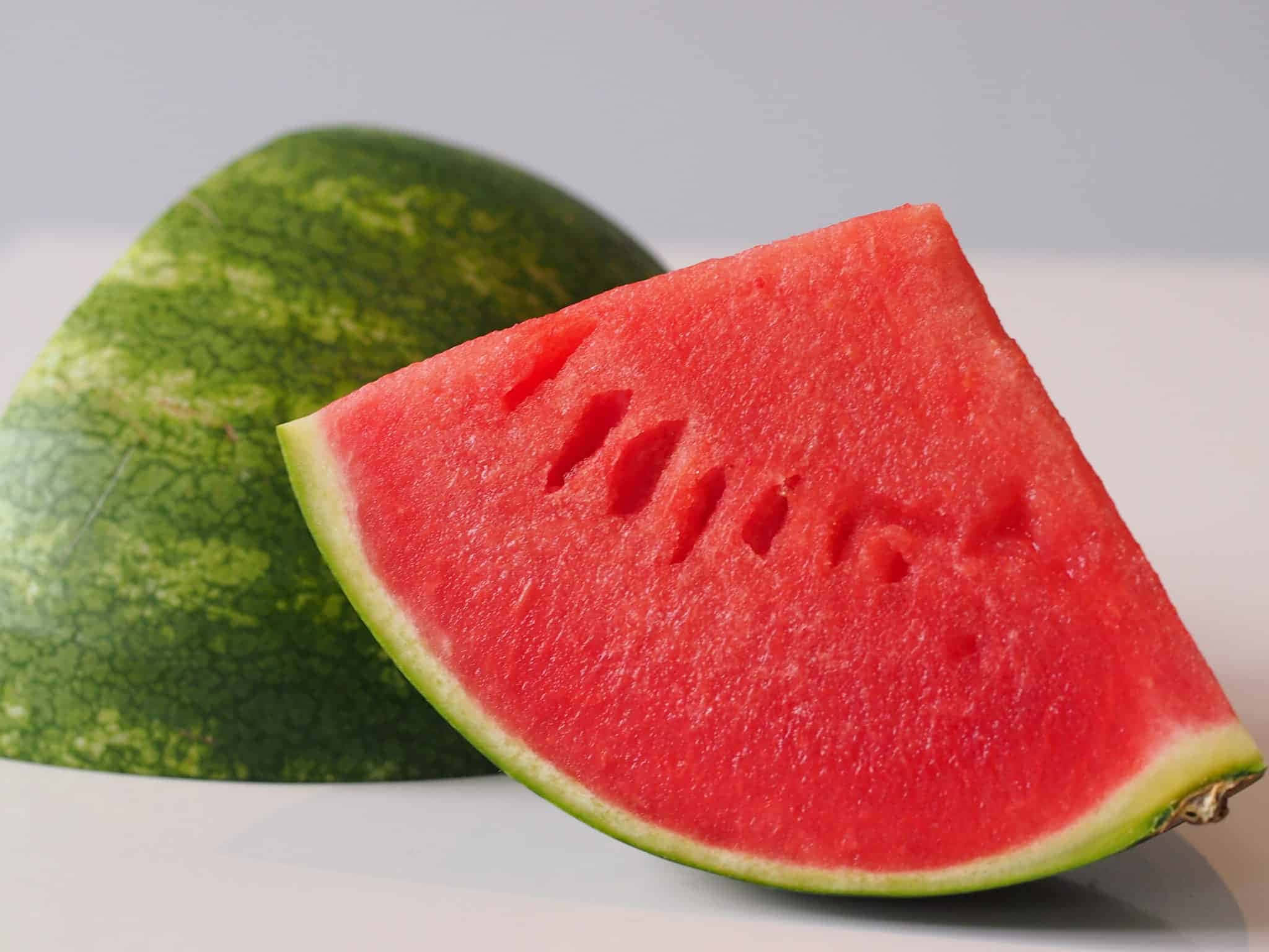 Watermelon 桌布 壁紙 — 一塊鮮紅多籽的西瓜片，旁邊放著一個完整的青皮西瓜，置於潔白的表面上。