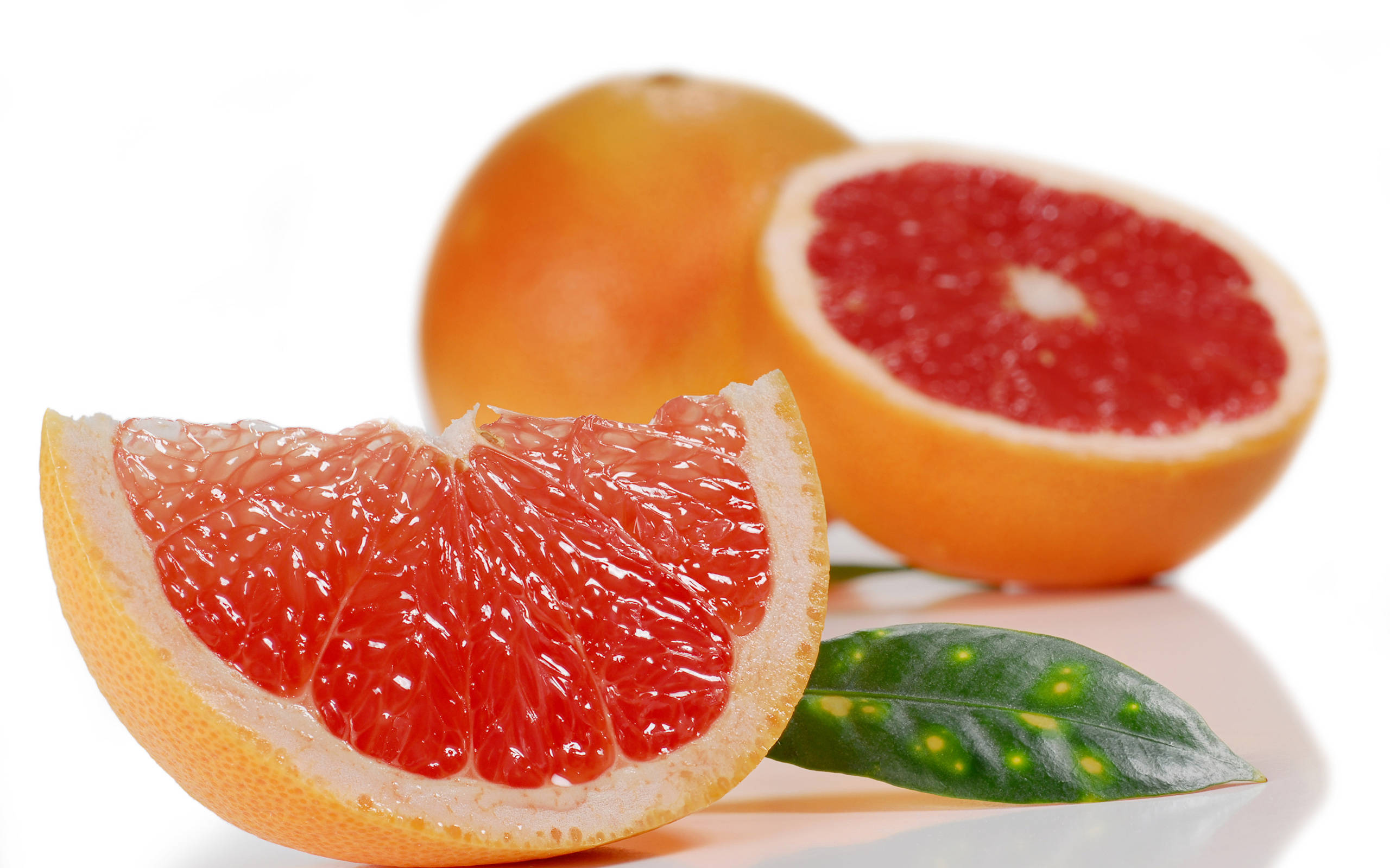 Fruits 桌布 壁紙 — 一塊鮮紅的葡萄柚片在潔白的表面上閃閃發光，旁邊有半個和整個葡萄柚，還有一片帶斑點的綠葉。