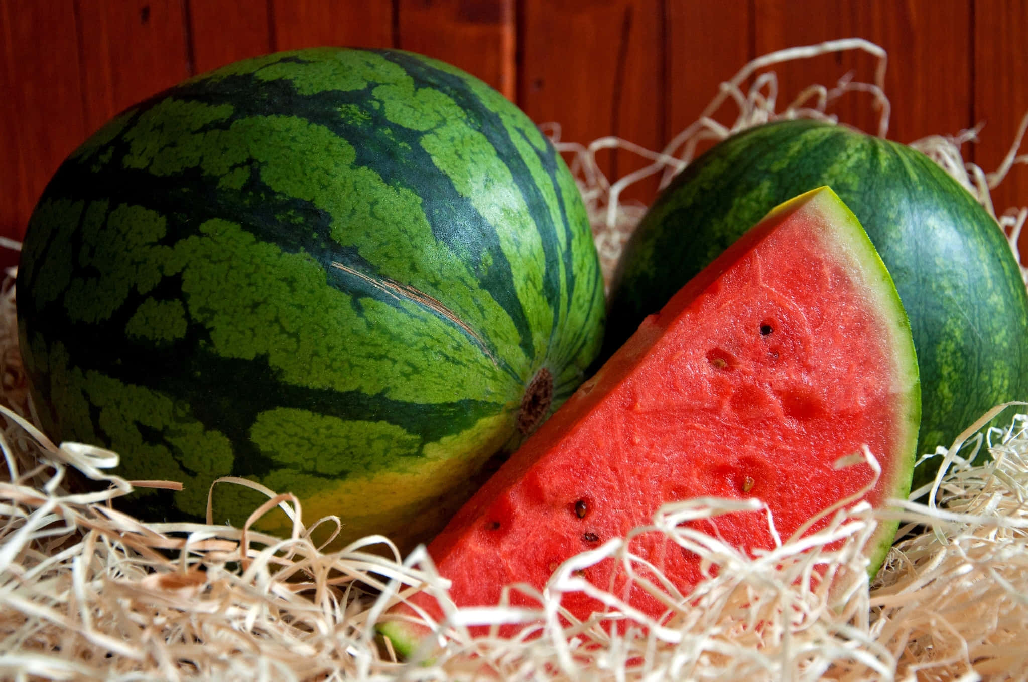 Watermelon 桌布 壁紙 — 一塊鮮紅色的西瓜切片，帶有黑色種子，放在木質背景前的稻草中，旁邊是完整的西瓜。
