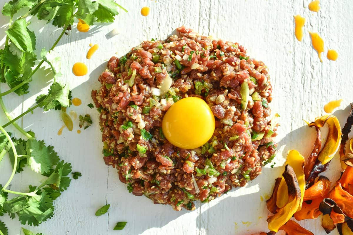meat 桌布 壁紙 — 一塊鮮豔的生牛肉他他，上面放著生蛋黃，放在白色木板上，周圍是新鮮的香菜和烤蔬菜。
