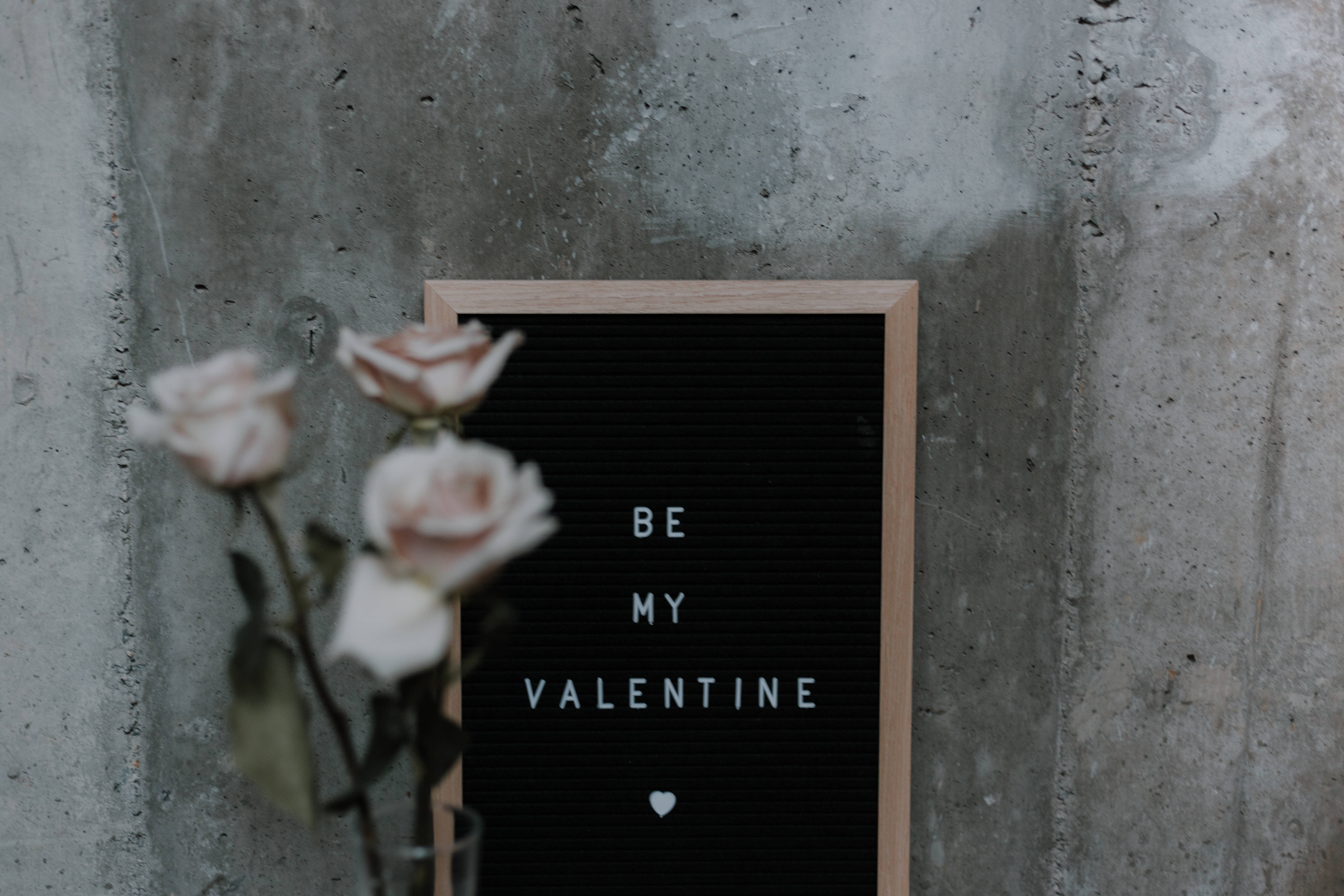 可愛情人節 桌布 壁紙 — 一塊黑色字母板，上面寫著「BE MY VALENTINE」和一個心形，靠著粗糙的混凝土牆，旁邊是玻璃花瓶中的柔軟粉紅玫瑰。