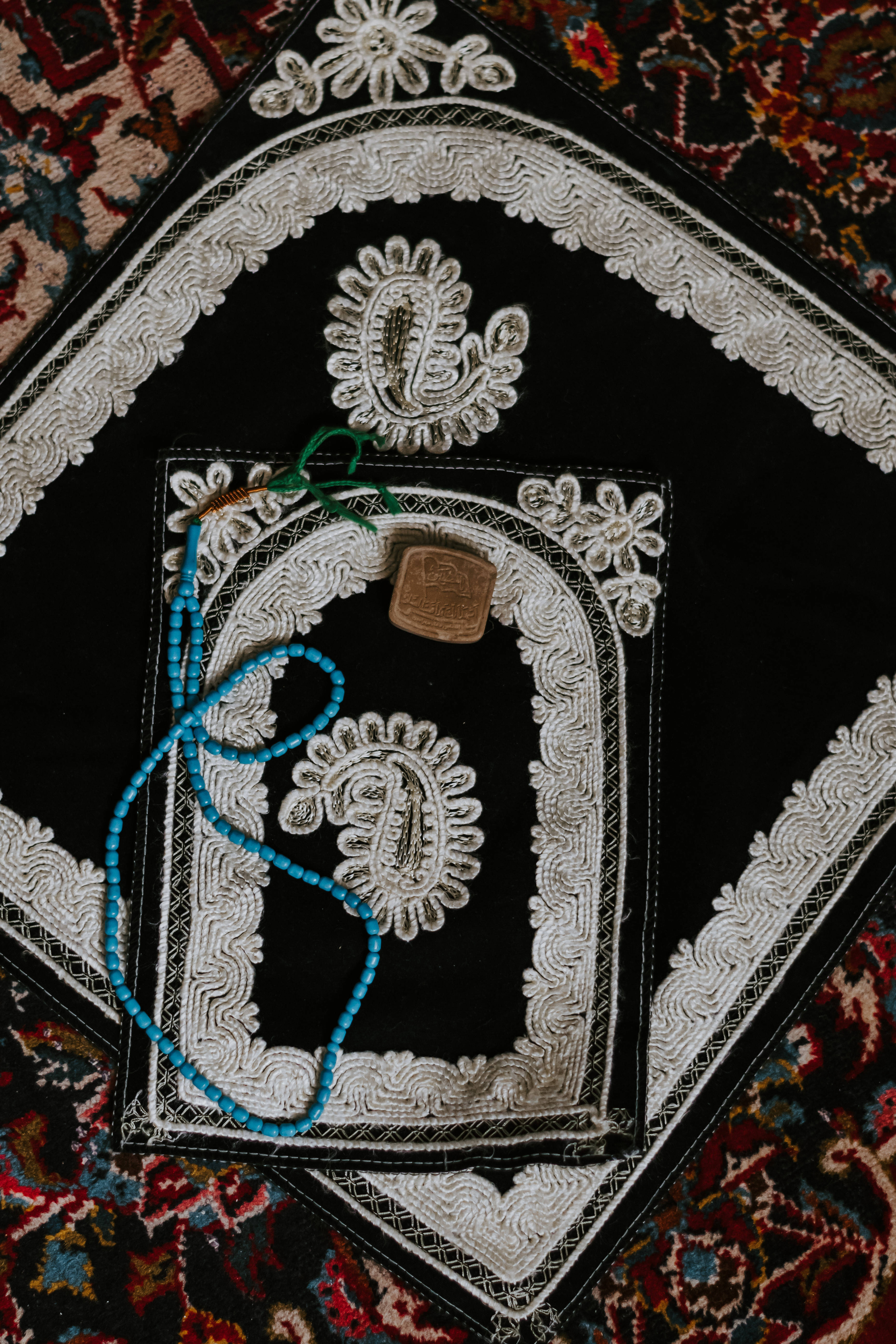 Ramadan 桌布 壁紙 — 一塊黑色祈禱地毯，帶有精緻的白色刺繡和一條藍色祈禱珠項鍊，放在色彩繽紛的圖案地毯上。