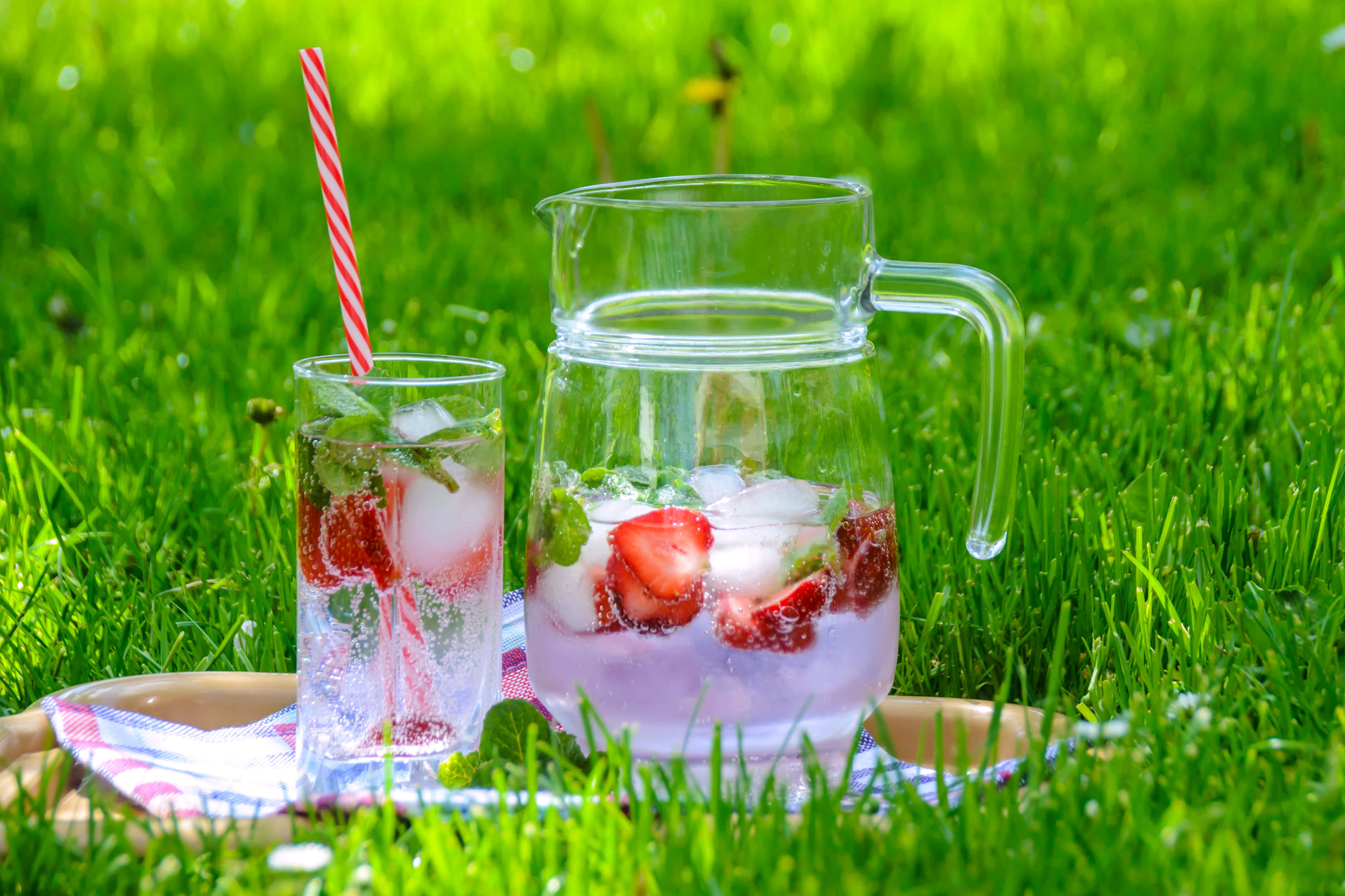 Summer Drinks 桌布 壁紙 — 一壺和一杯清涼的草莓薄荷檸檬水，加冰和條紋吸管，放在翠綠草地上的格子托盤上。