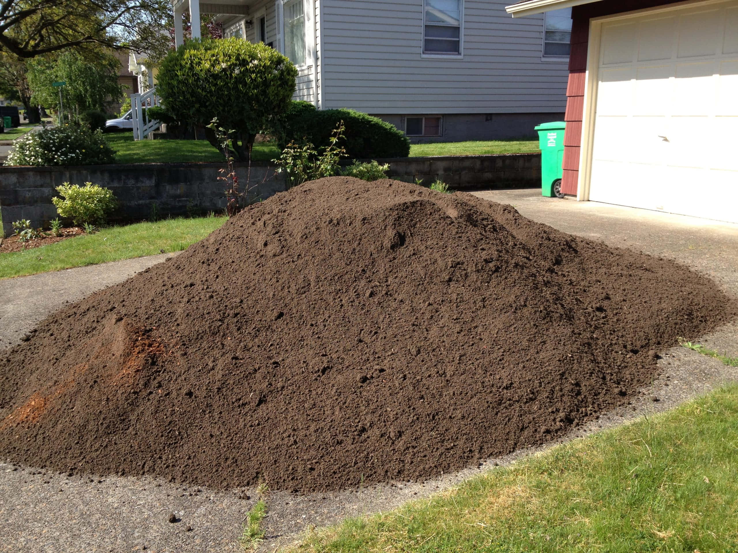 Dirt 桌布 壁紙 — 一大塊肥沃的深色泥土堆放在郊區房屋旁邊的車道上，房屋周圍有綠草和灌木，在明亮的日光下。
