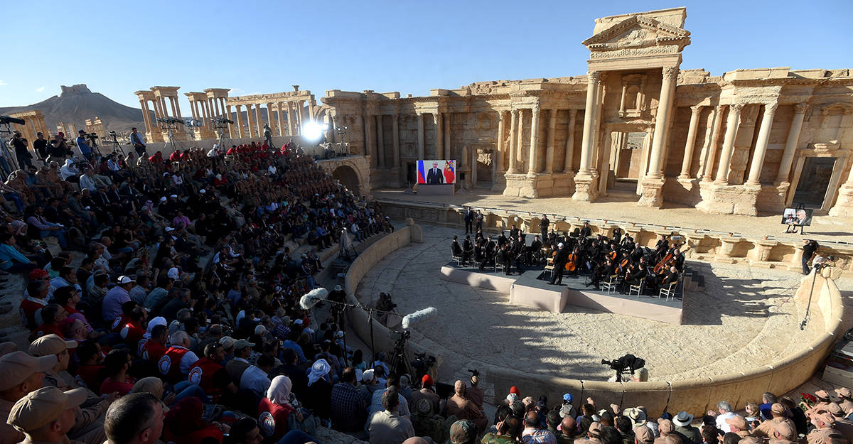 Syria 桌布 壁紙 — 一大群人聚集在古老的石造圓形劇場，管弦樂團在明亮的日光下演奏。