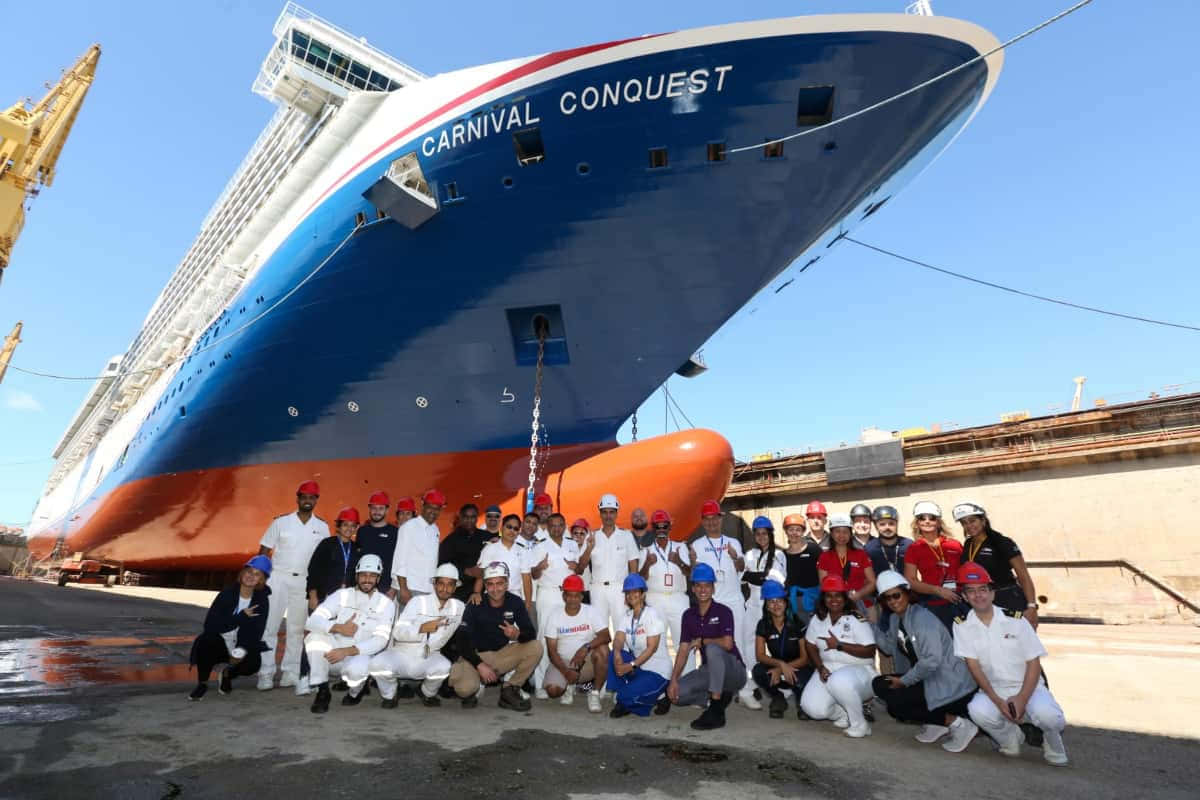 Carnival Conquest 桌布 壁紙 — 一大群戴著安全帽的工人在晴朗藍天下自豪地站在船塢停泊的巨大藍橙色嘉年華征服號郵輪前合影。