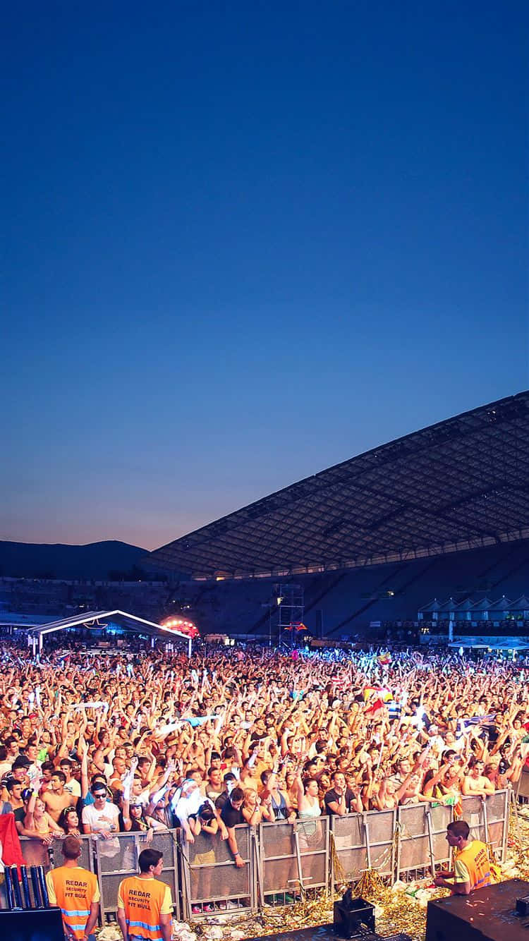 concert 桌布 壁紙 — 一大群演唱會觀眾在暮色天空下的體育場中舉起雙手，舞台燈光閃爍。