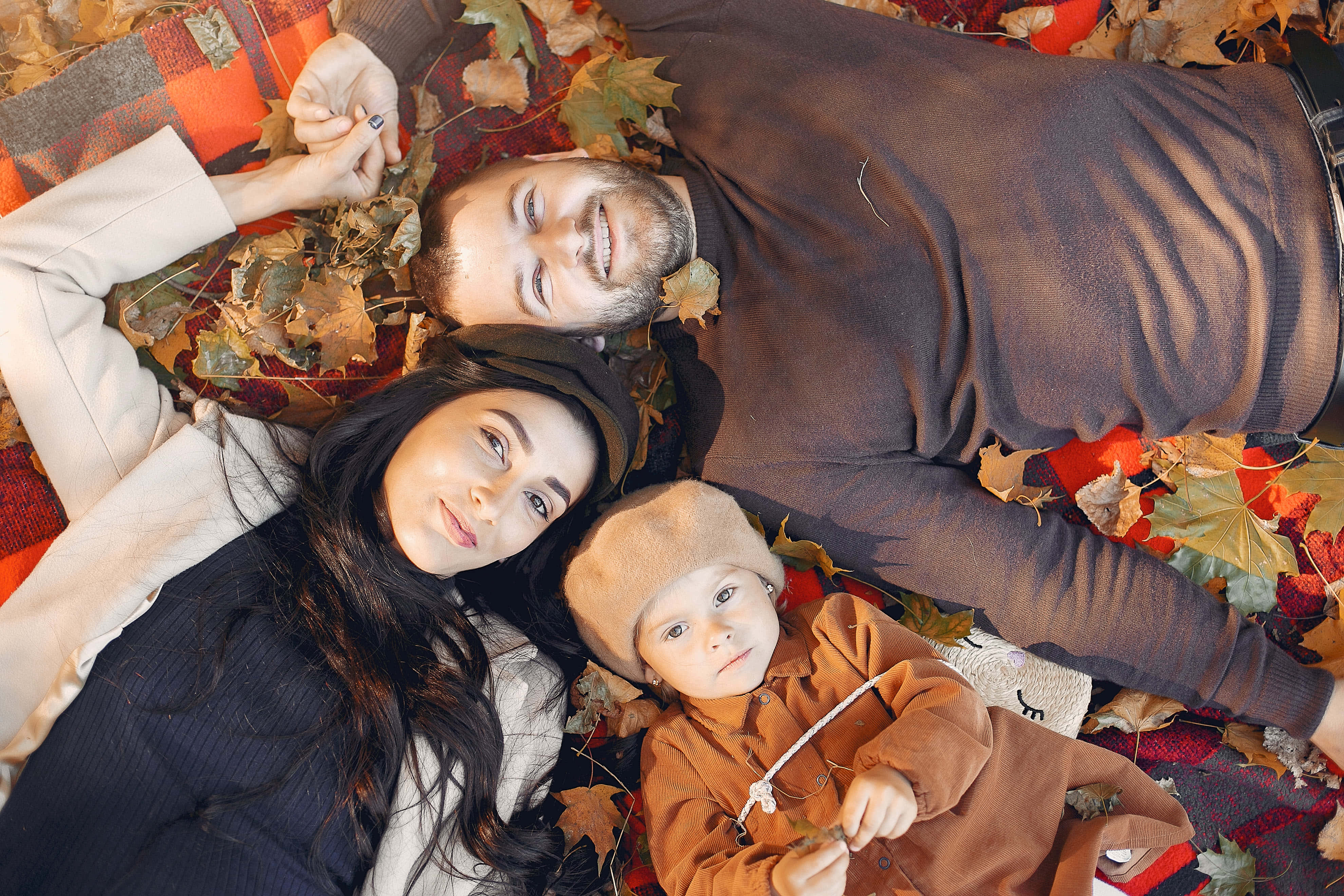 Fall Family 桌布 壁紙 — 一家三口在柔和日光下，一起躺在格紋毯上，周圍是鮮豔的秋葉。