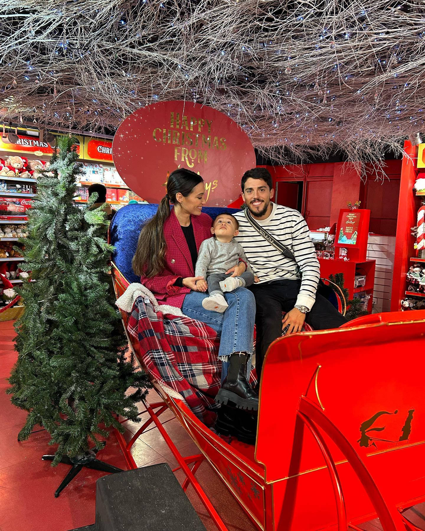 Celebrity 桌布 壁紙 — 一家三口在紅色雪橇上微笑，背景是閃爍的聖誕樹和「Happy Christmas」標誌，充滿節日氣氛的商店。
