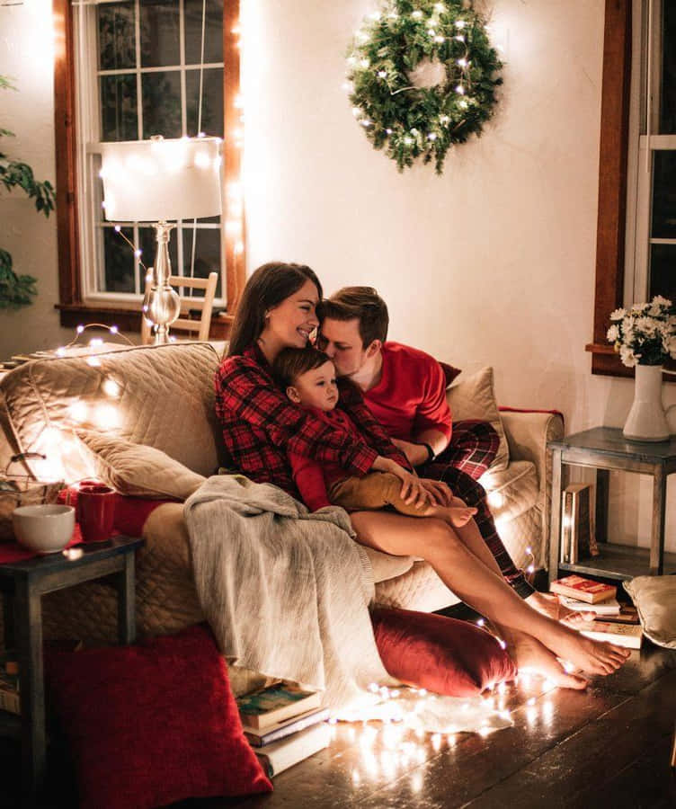 Family Christmas 桌布 壁紙 — 一家三口在舒適的客廳中，被閃爍的童話燈和節日裝飾包圍，溫暖地依偎在沙發上。
