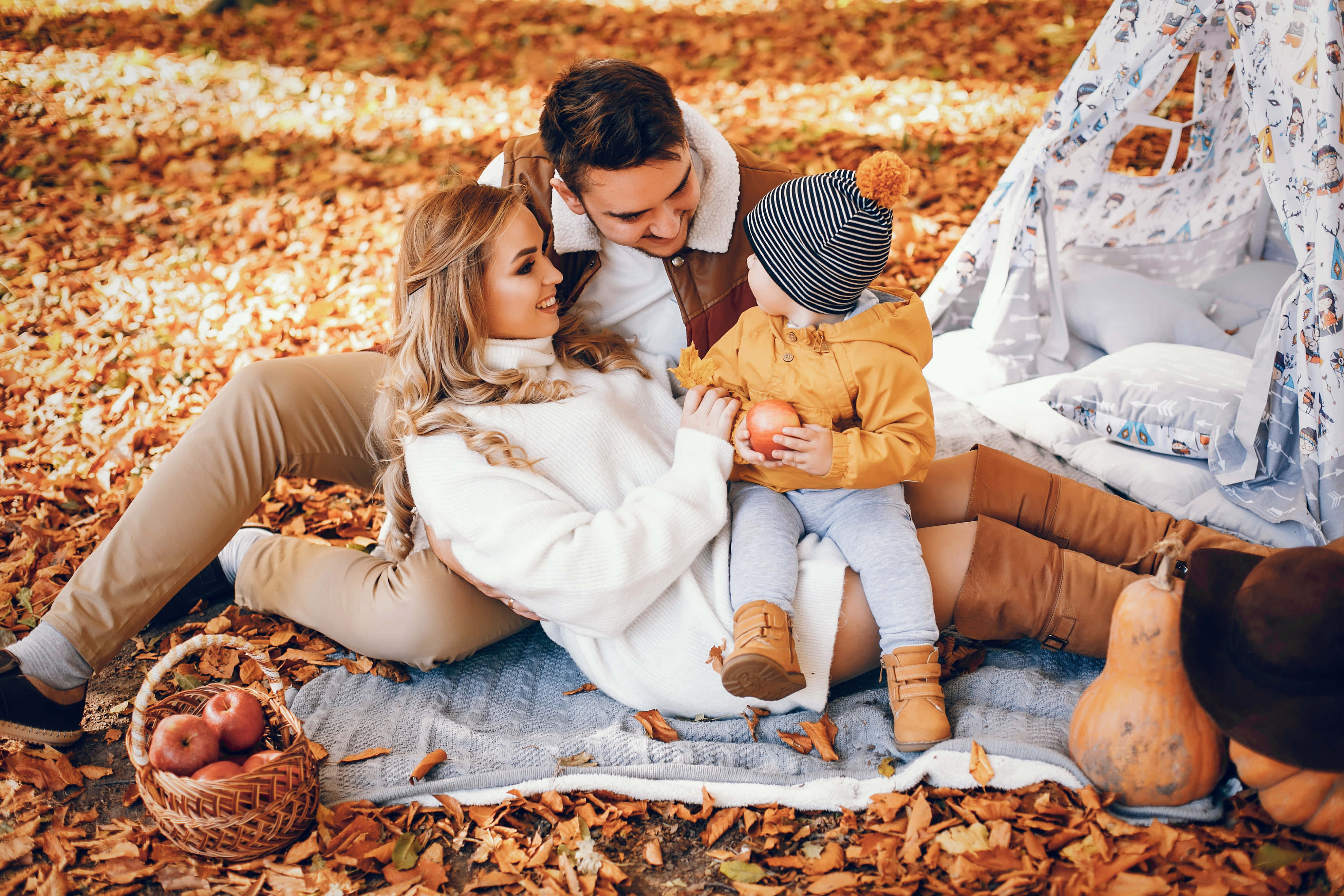 Fall Family 桌布 壁紙 — 一家三口在金色樹葉的天篷下共享舒適的秋季野餐，附近有南瓜和柳條籃。