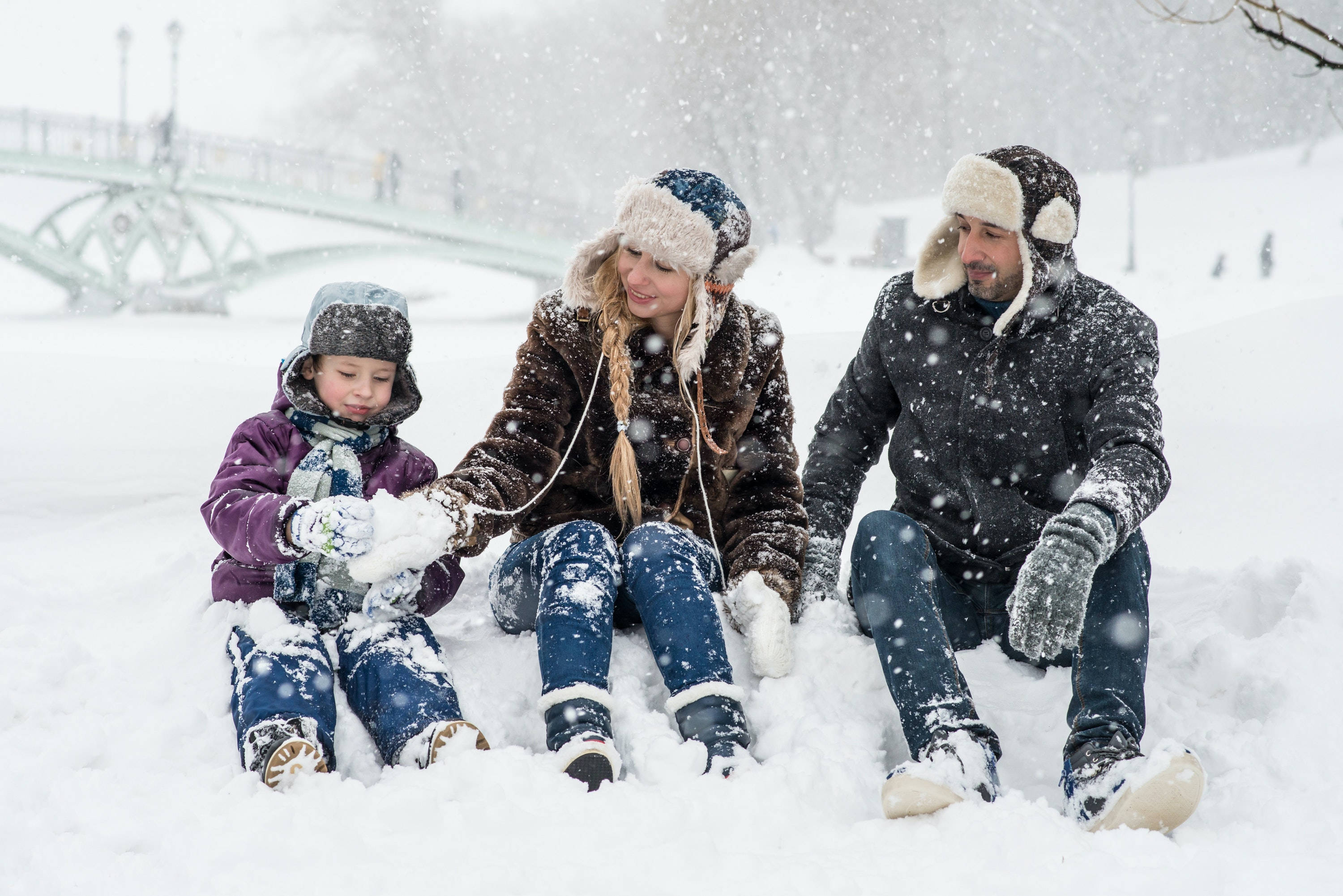 Family 桌布 壁紙 — 一家三口坐在厚厚的雪中，在飄落的雪花下一起堆雪球，背景可見一座橋。