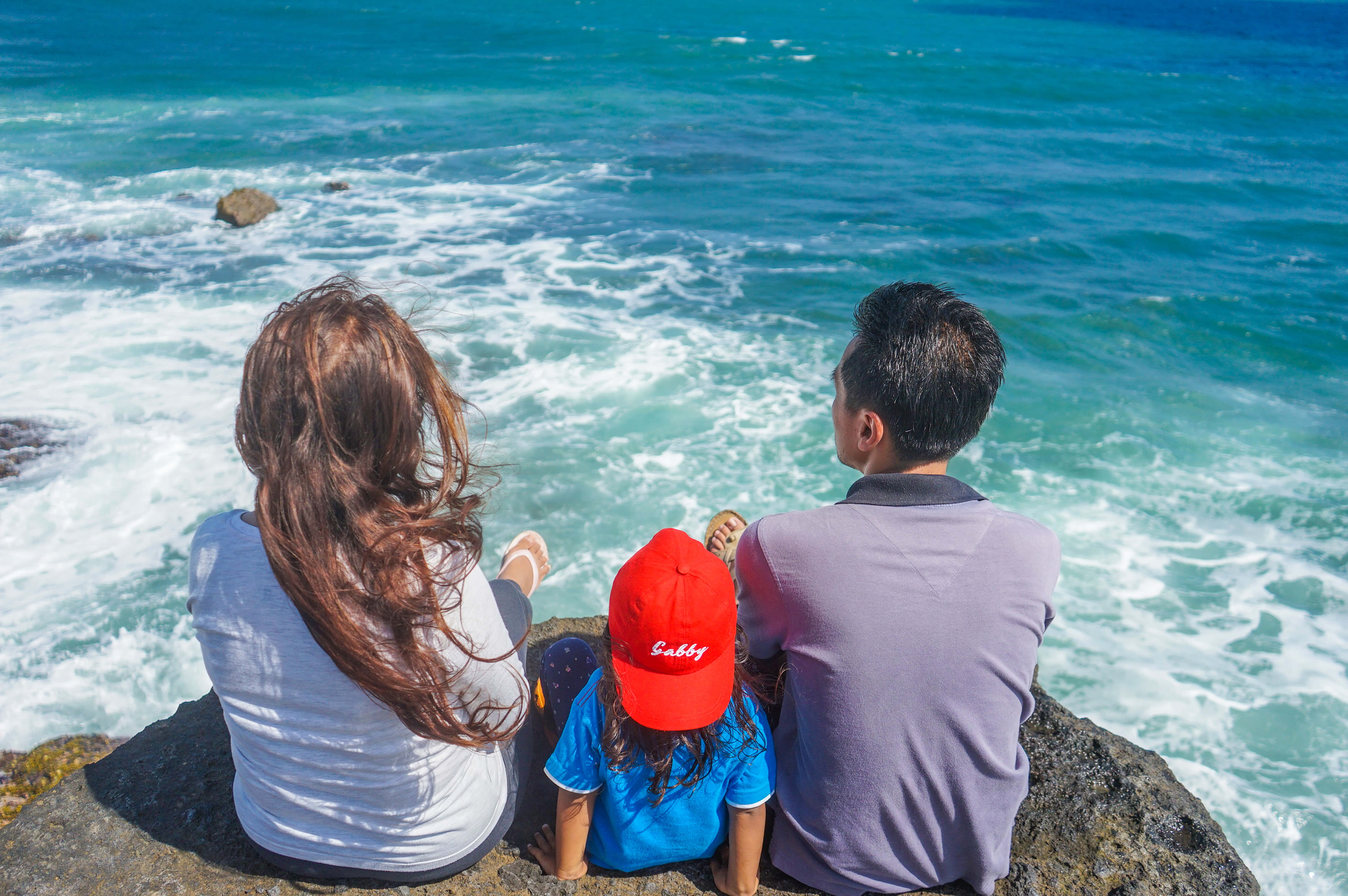 Summer Vacation 桌布 壁紙 — 一家三口坐在岩石懸崖上，俯瞰下方在明亮陽光下拍打的碧綠海浪。