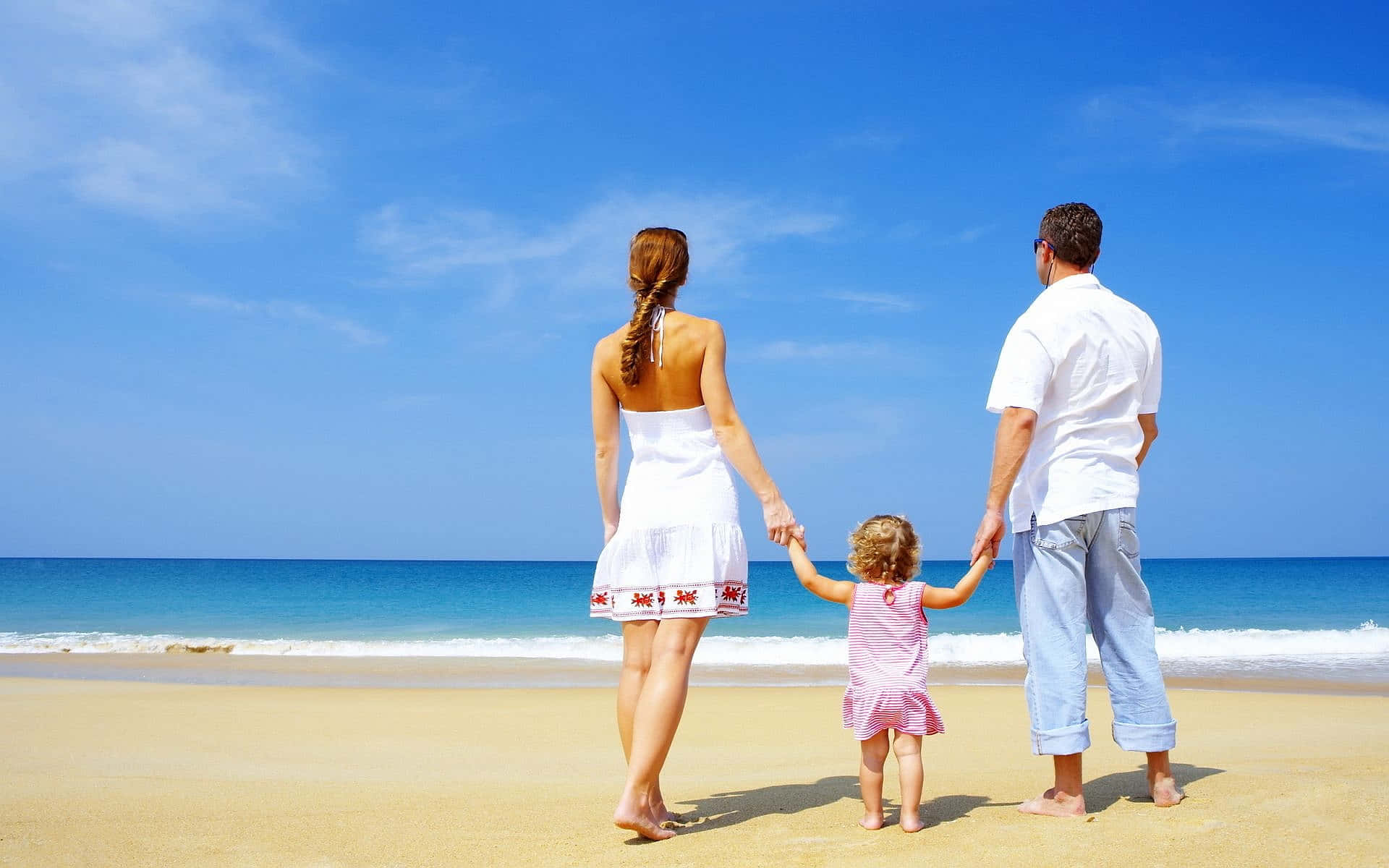 Beach Walk 桌布 壁紙 — 一家三口手牽手沿著陽光明媚的海灘散步，海面碧綠，天空湛藍。