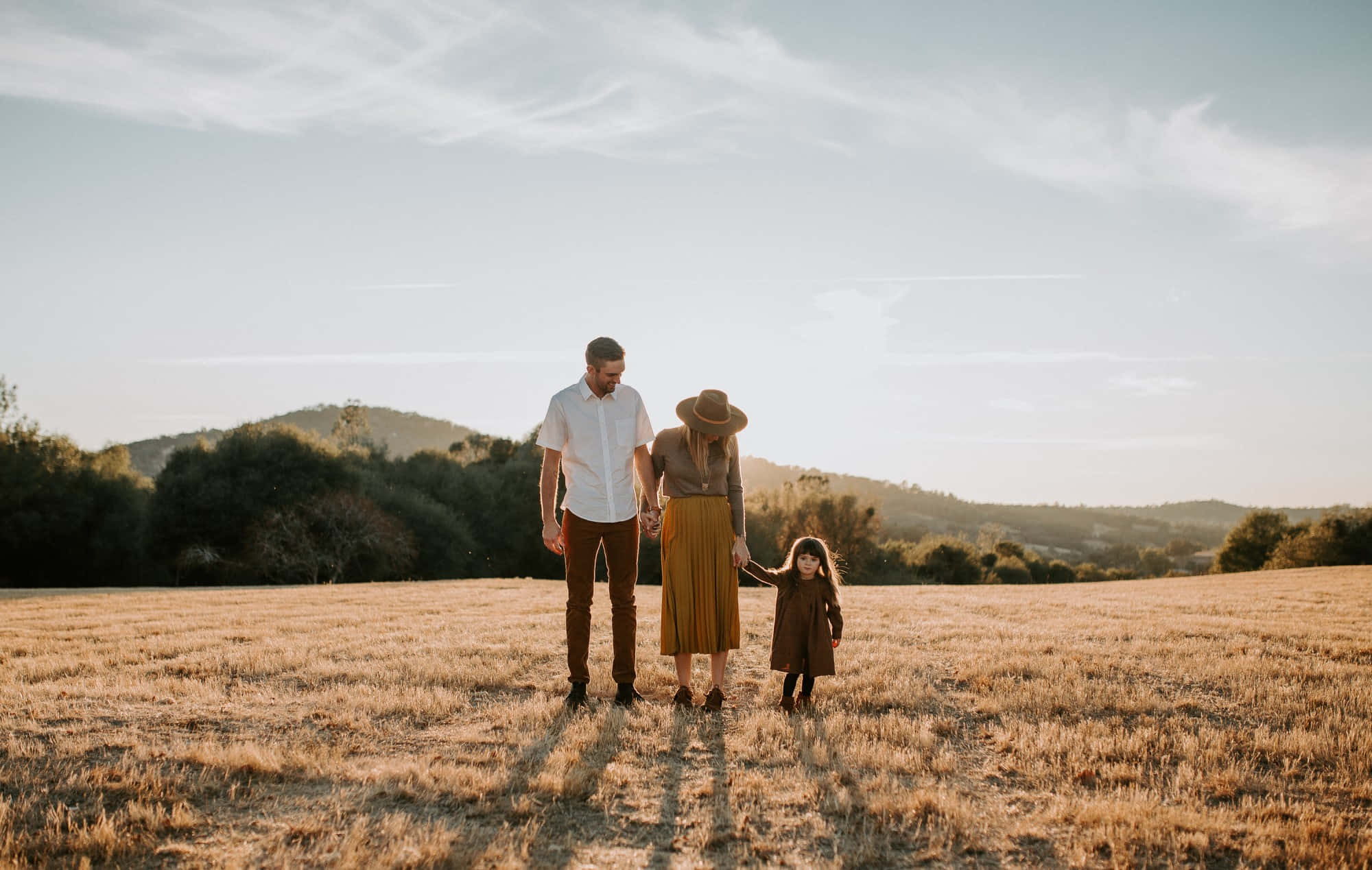 Fall Family 桌布 壁紙 — 一家三口手牽手走過日落時分的金色田野，背景是連綿起伏的山丘。