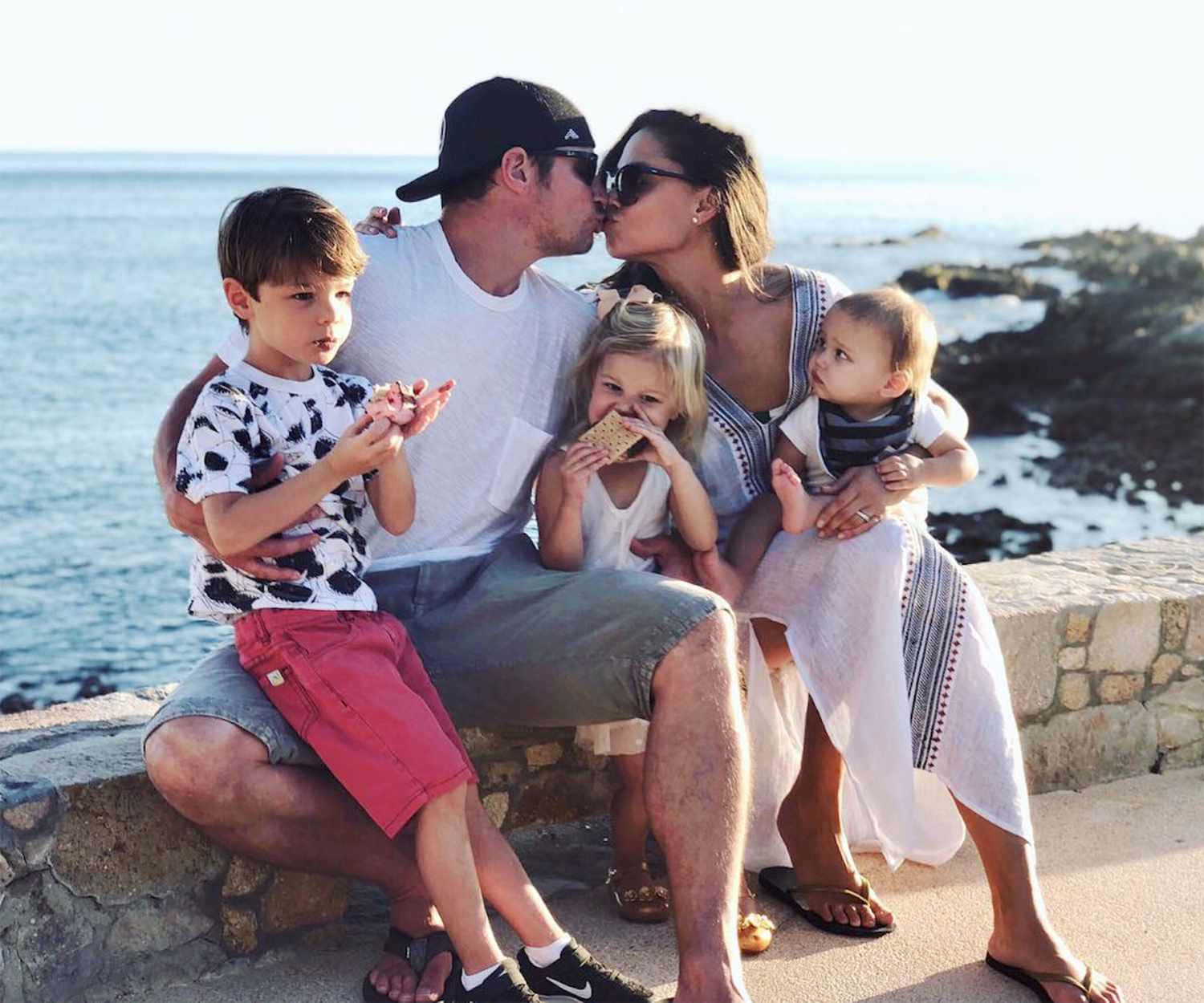 Family 桌布 壁紙 — 一家五口在陽光照耀的石牆上分享溫馨的海邊時光，海浪在附近拍打。